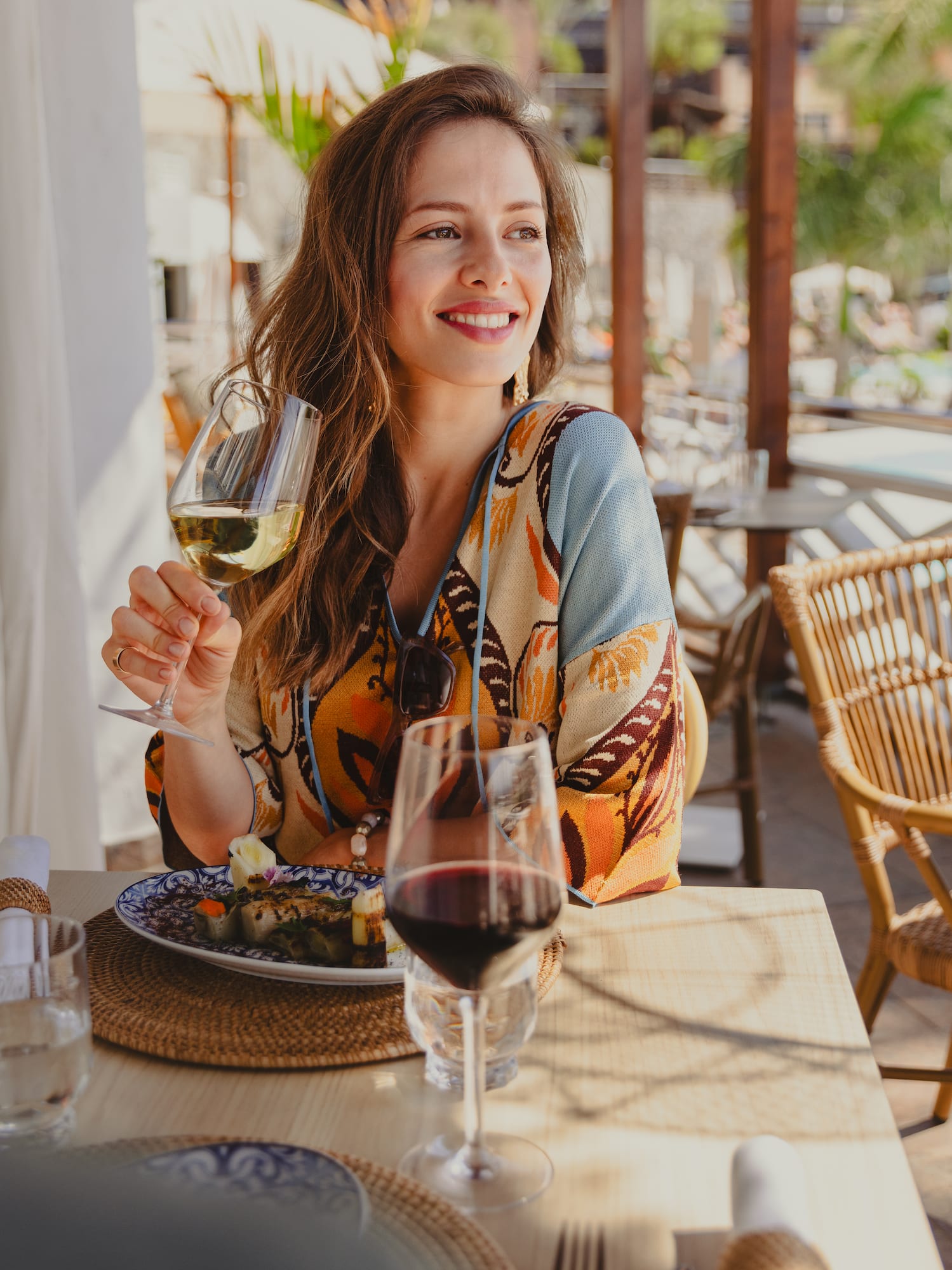 Smiling woman holding white wine, dining outdoors at a tropical resort restaurant.
