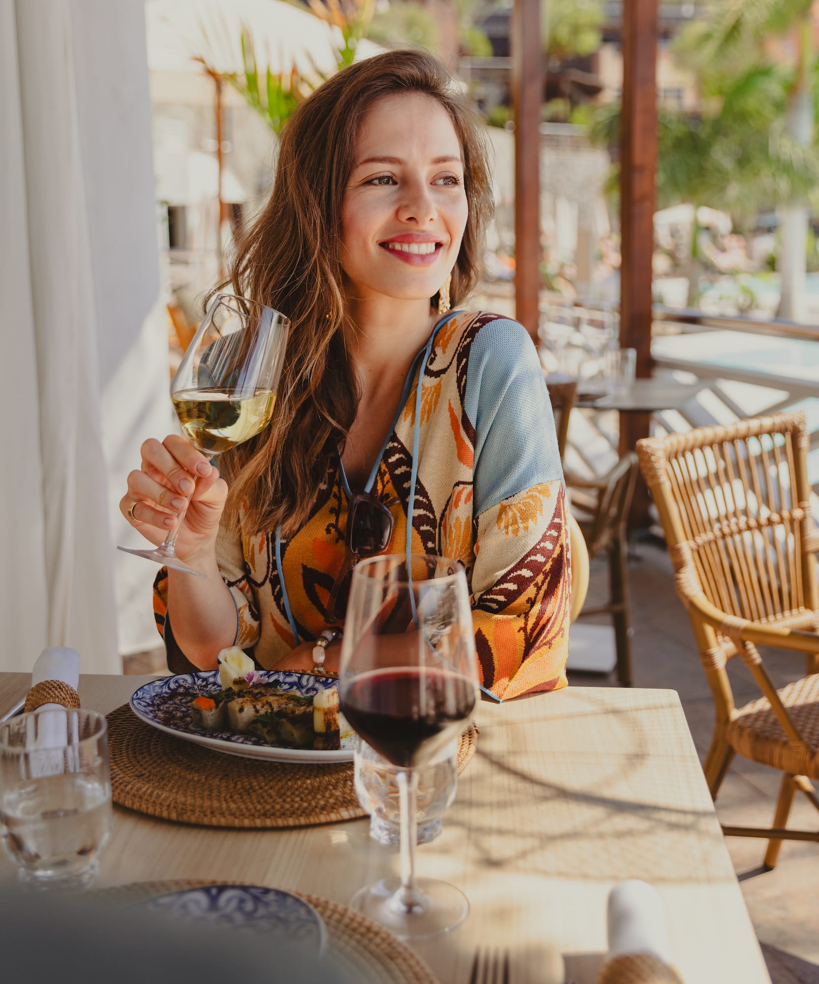 Smiling woman holding white wine, dining outdoors at a tropical resort restaurant.