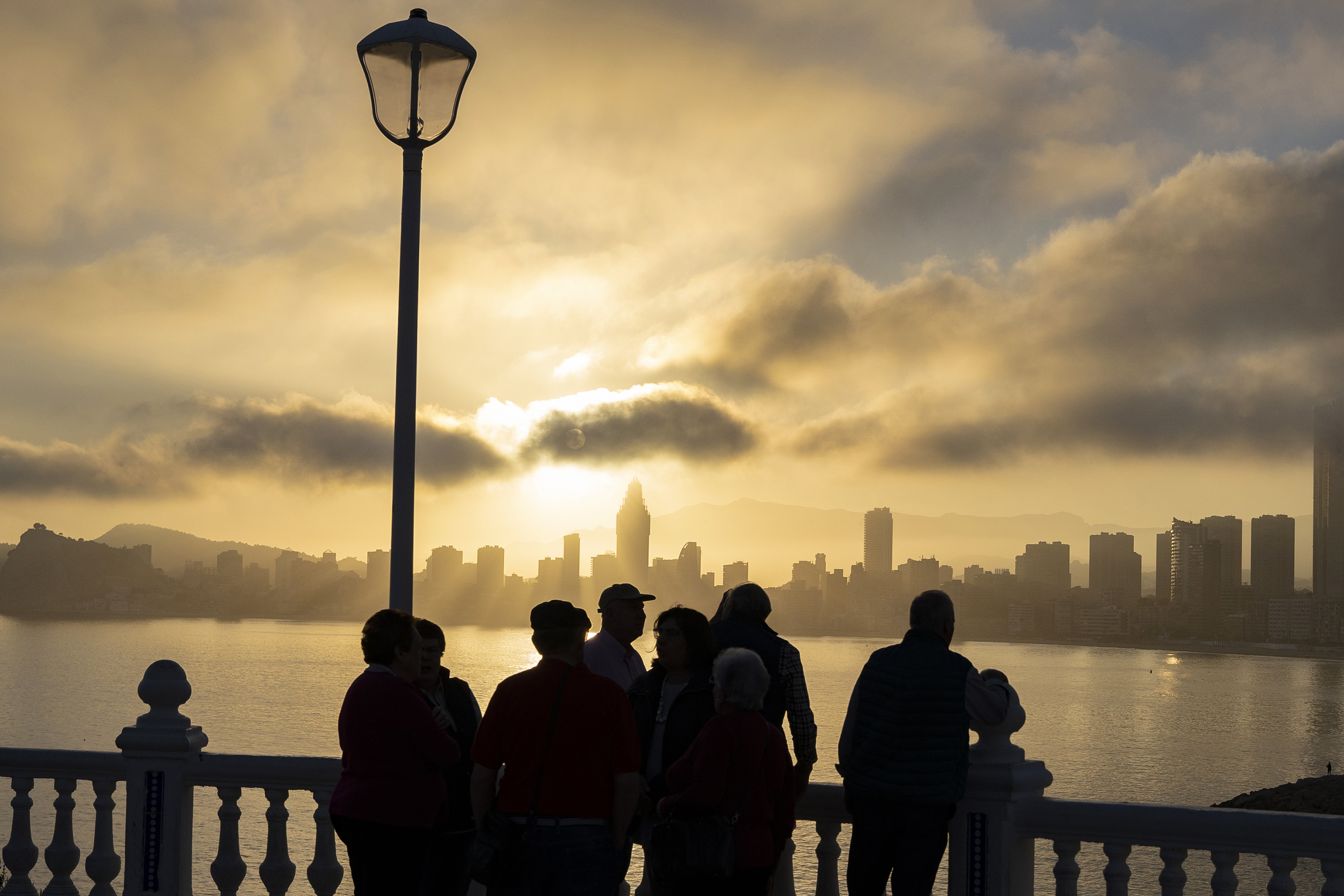Silhouetted people and city skyline across bay at golden sunrise.