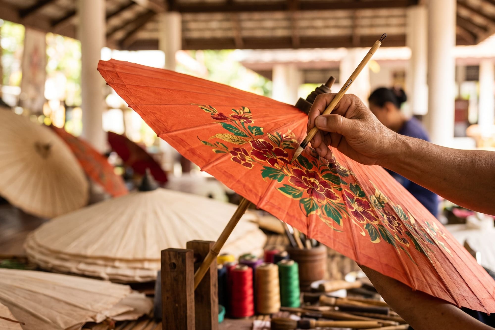 Thai artisan hand paints floral design on an orange paper umbrella.