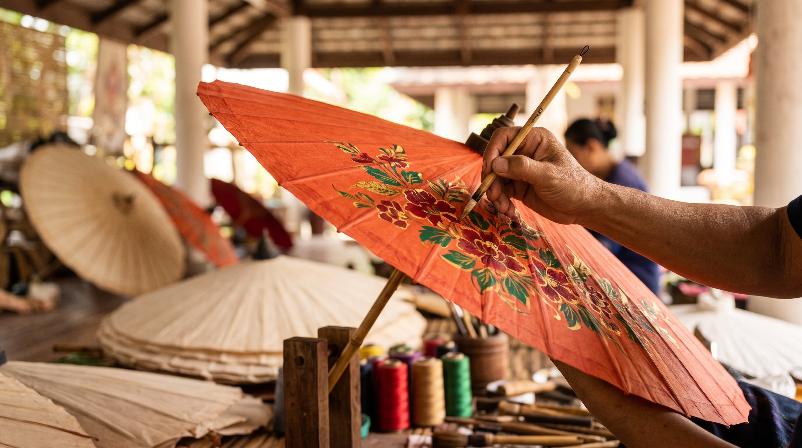 Thai artisan hand paints floral design on an orange paper umbrella.