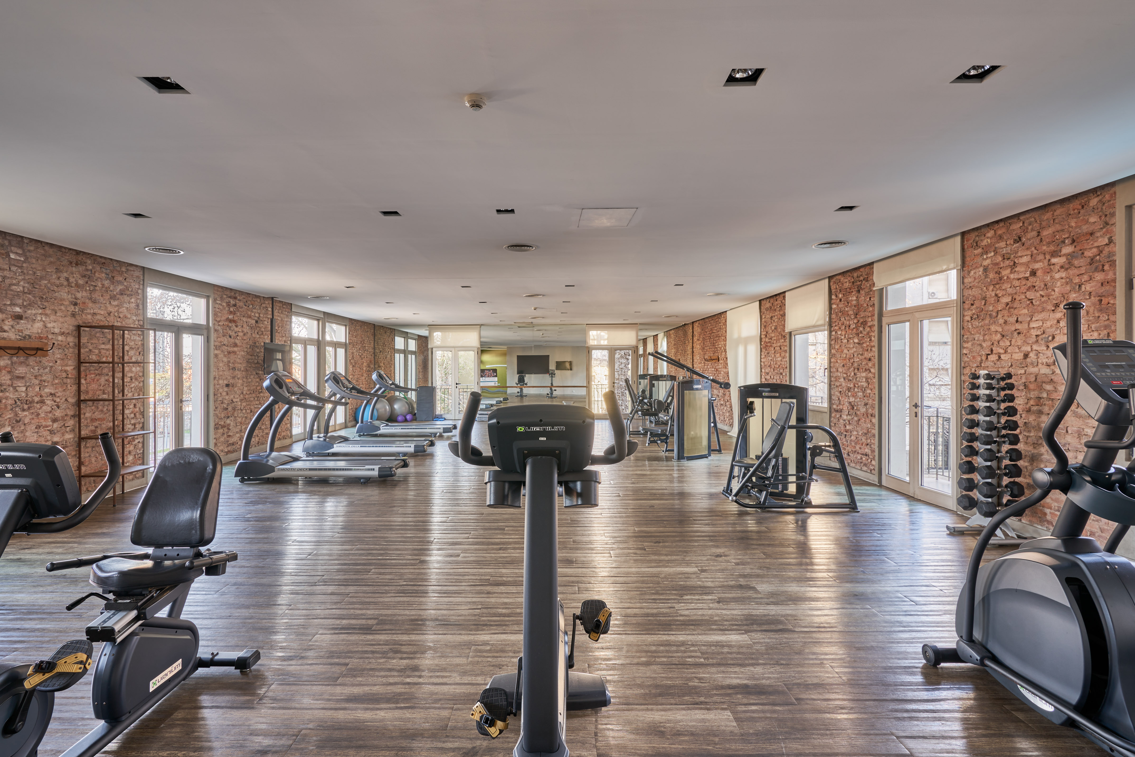 Modern gym with exercise equipment, brick walls, wood floor, and large windows.