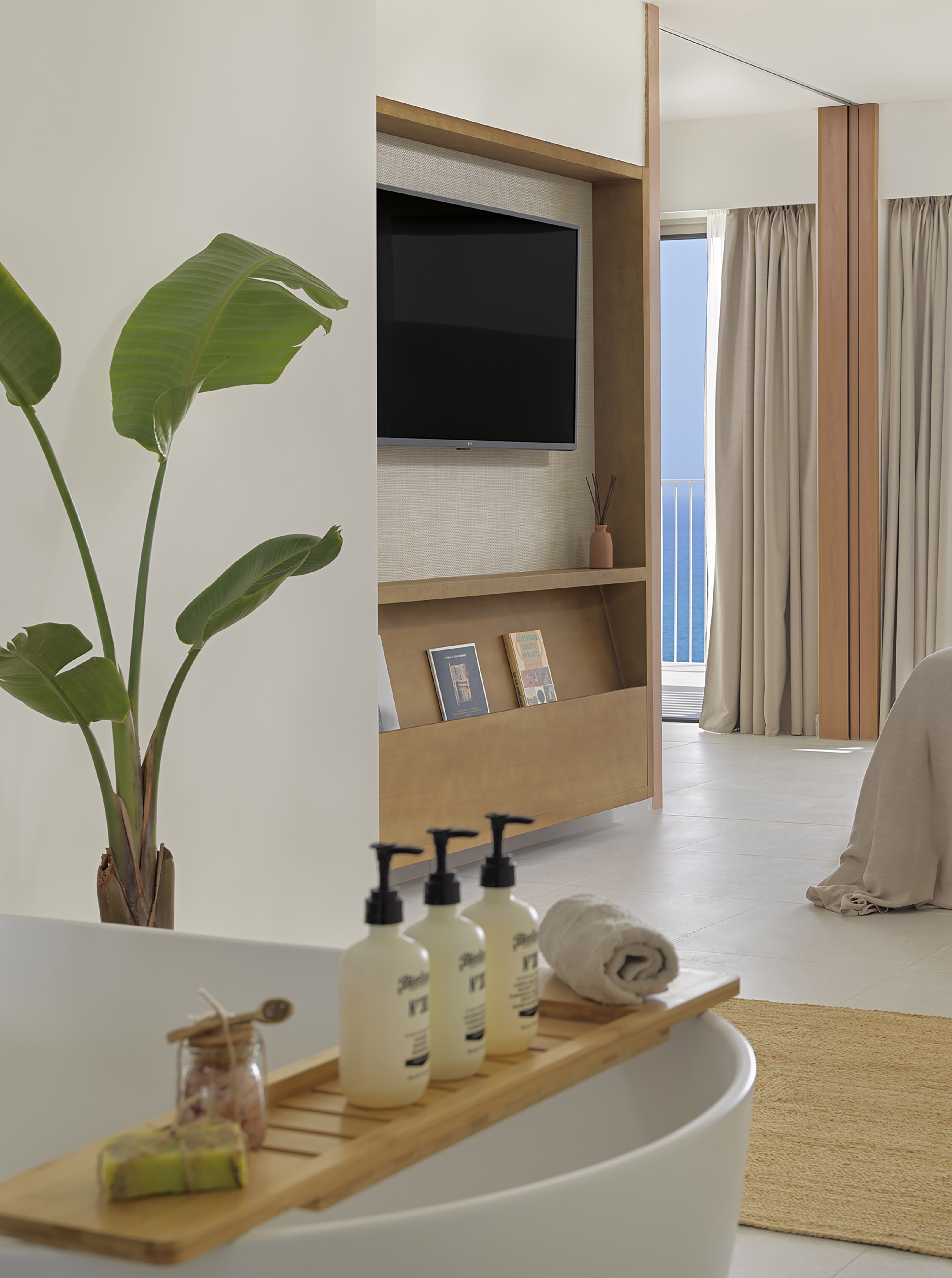 Modern hotel room with bathtub caddy, tropical plant, TV, and ocean view.
