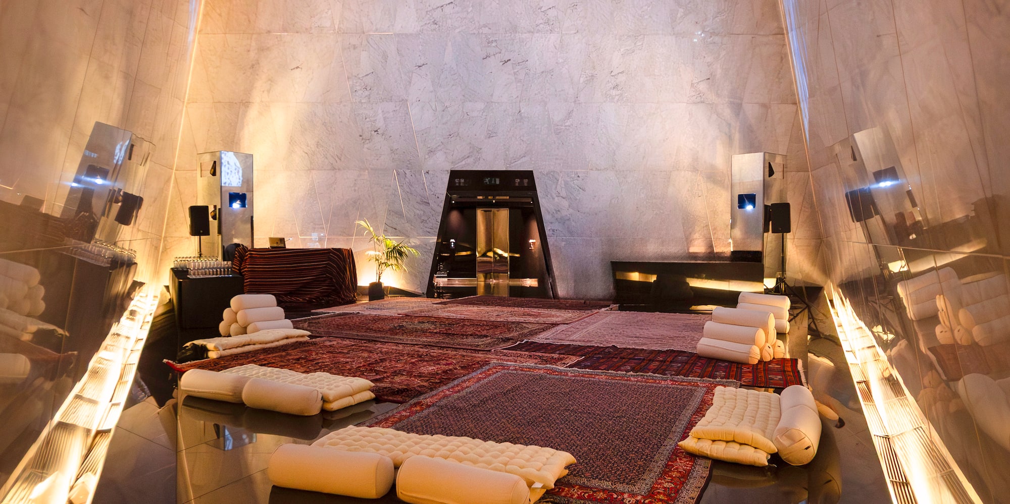 Modern lounge with high marble walls, reflective floor, oriental rugs, and cushions.