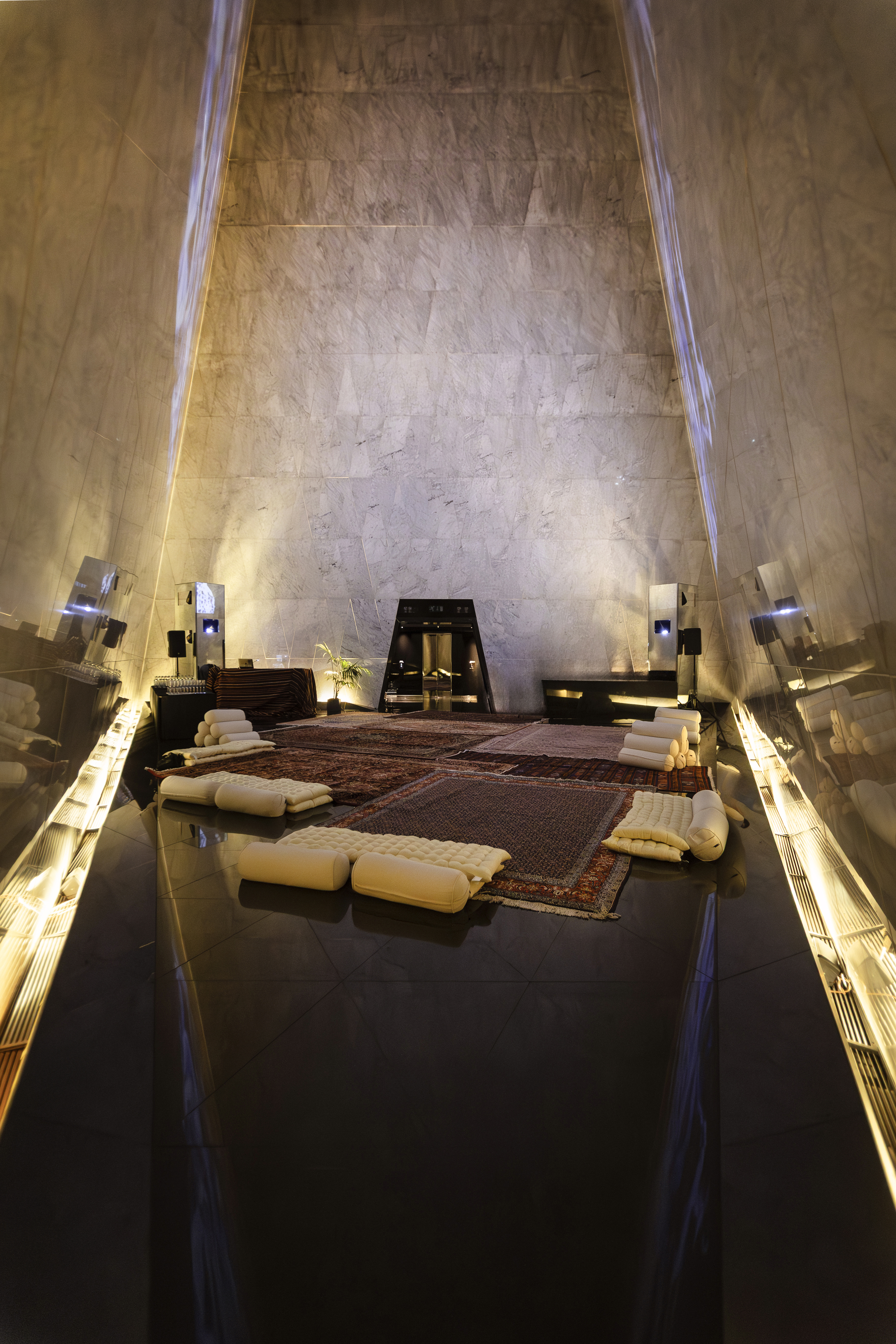 Modern lounge with high marble walls, reflective floor, oriental rugs, and cushions.