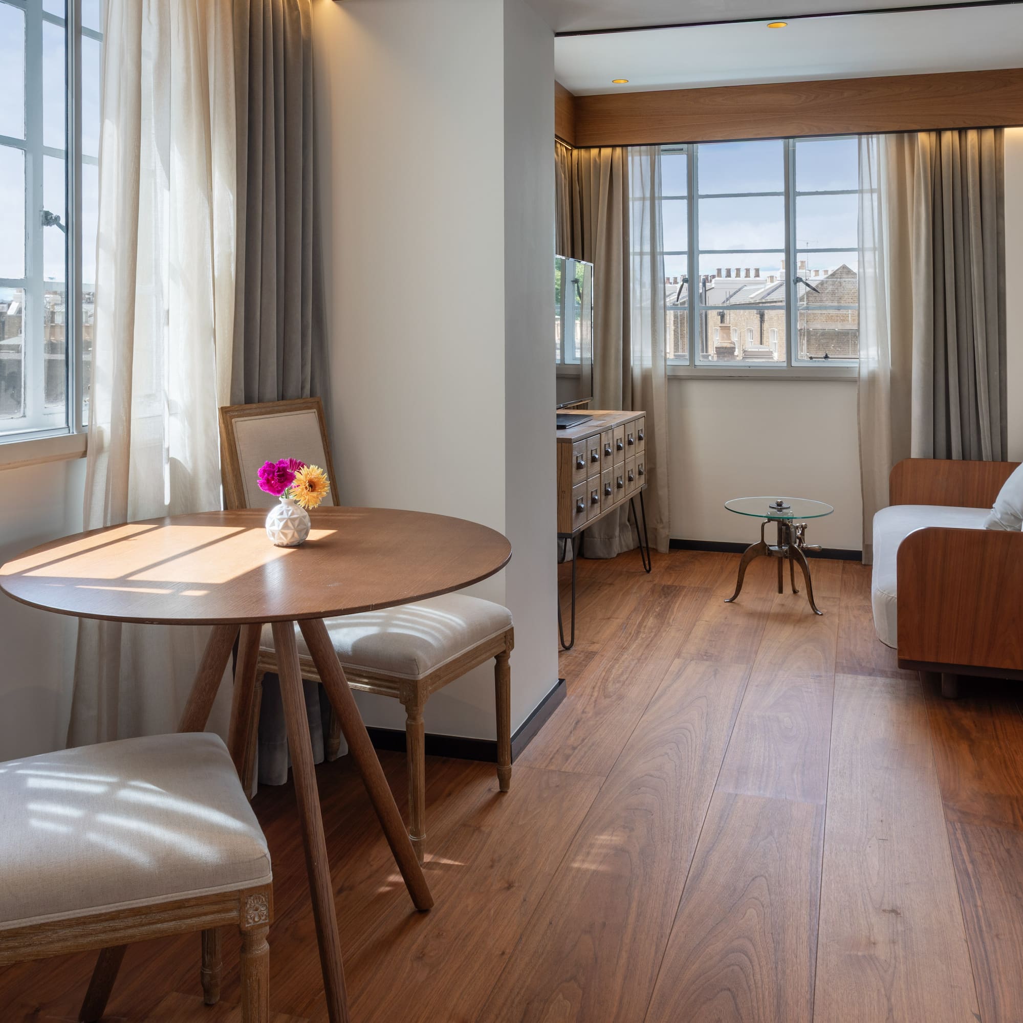 Modern hotel suite with dining table, sofa, wood floor, and sunlit windows.