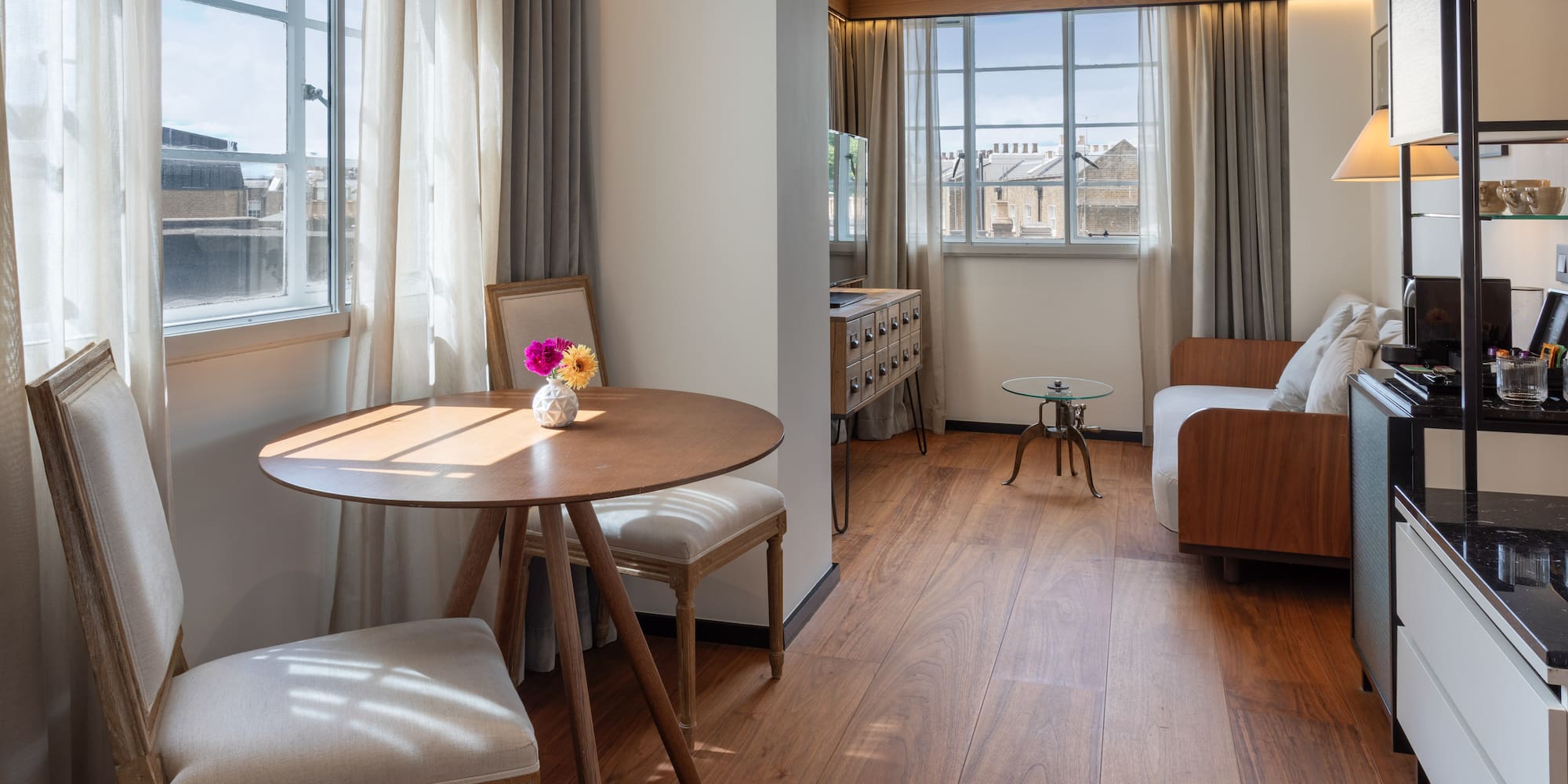 Modern hotel suite with dining table, sofa, wood floor, and sunlit windows.