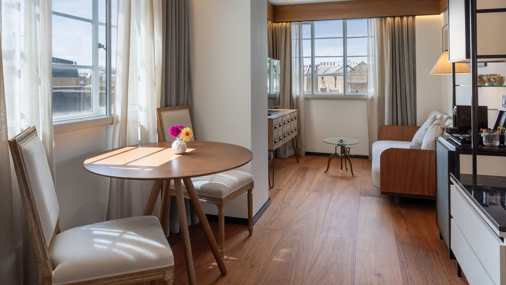 Modern hotel suite with dining table, sofa, wood floor, and sunlit windows.