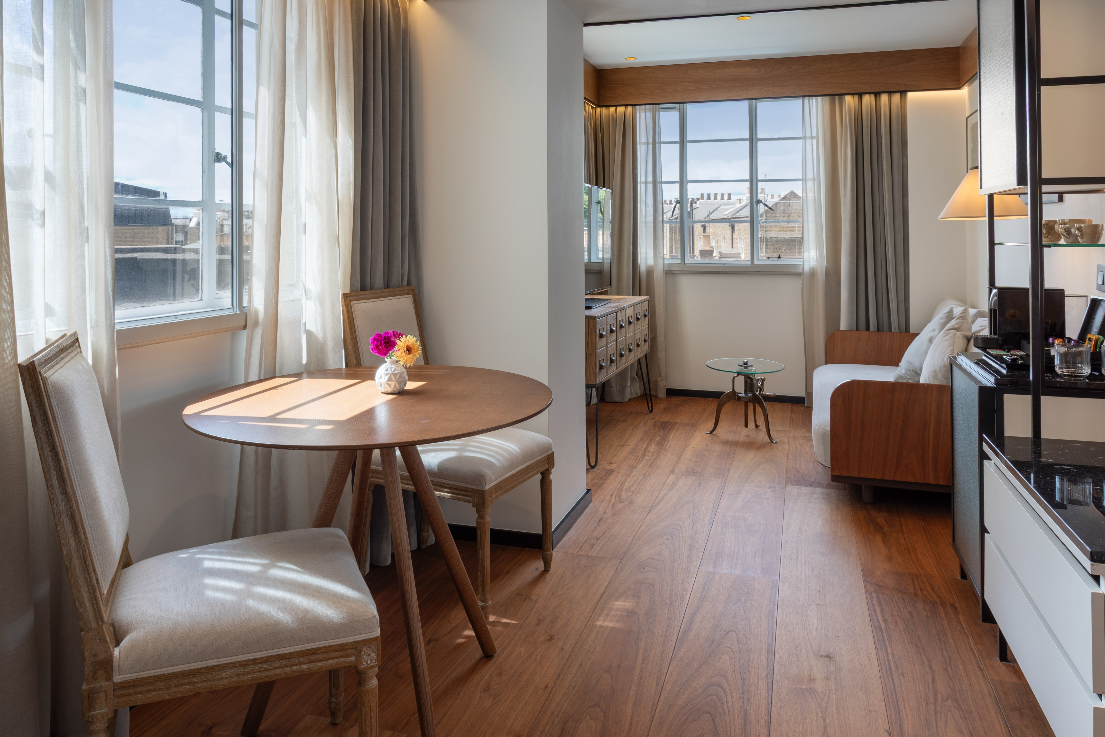 Modern hotel suite with dining table, sofa, wood floor, and sunlit windows.