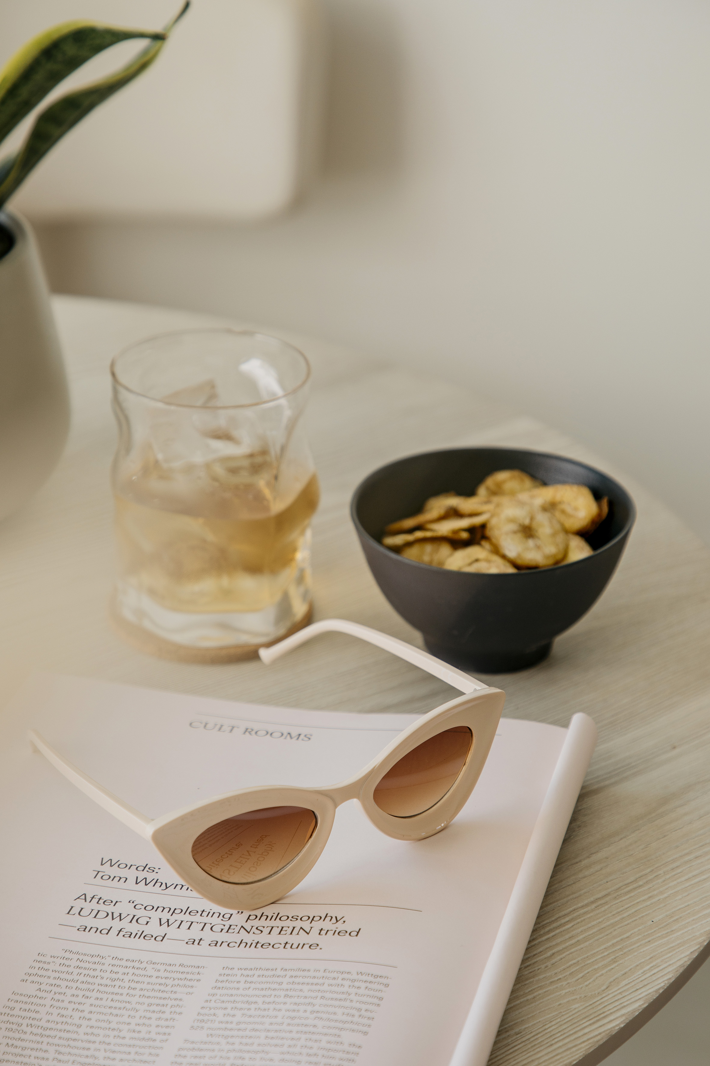 Cat-eye sunglasses, iced drink, banana chips, and open magazine on wooden table.