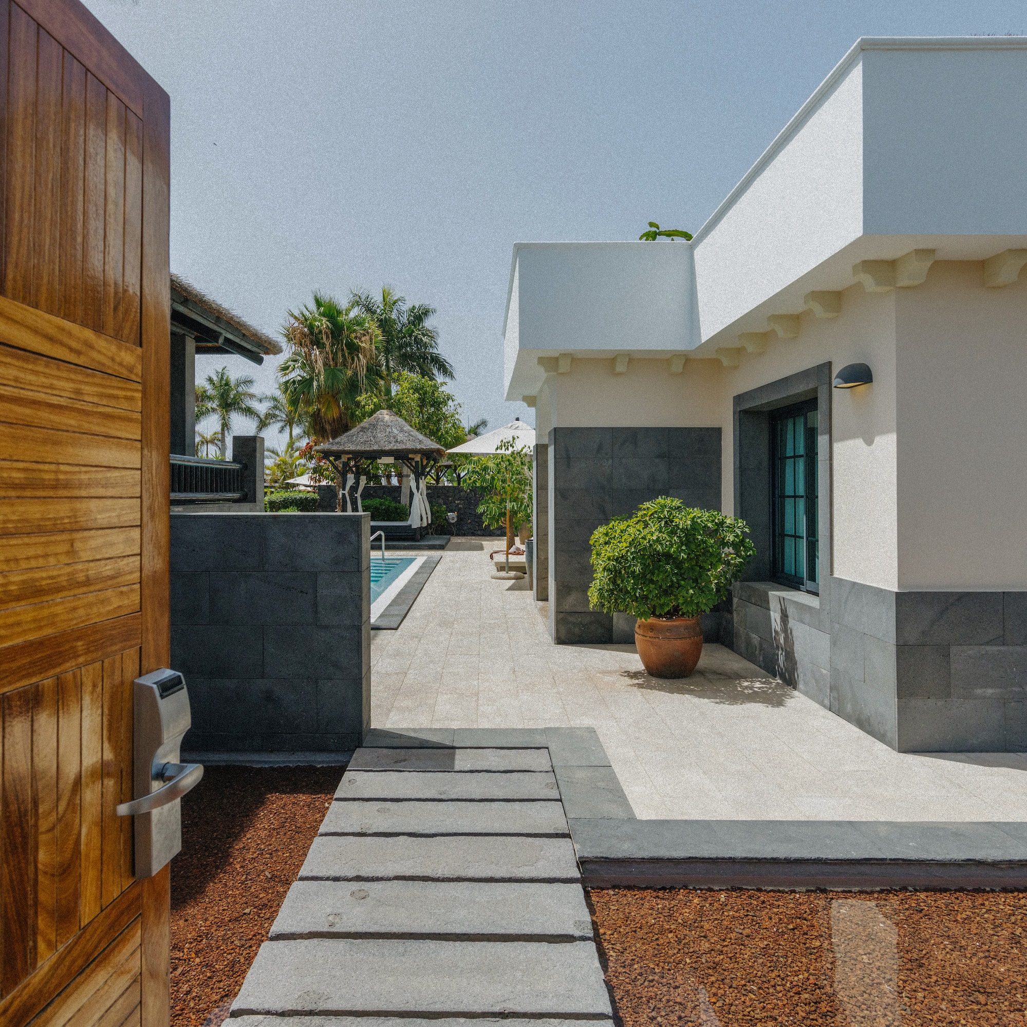 Open wooden gate to modern villa, stone path, outdoor pool, tropical resort.