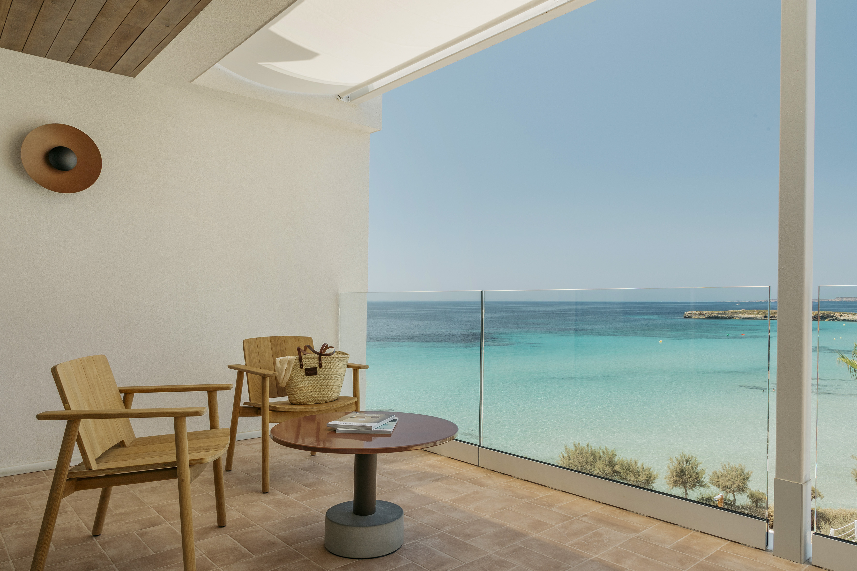 Sunny hotel balcony with wooden chairs, coffee table, and turquoise ocean view.