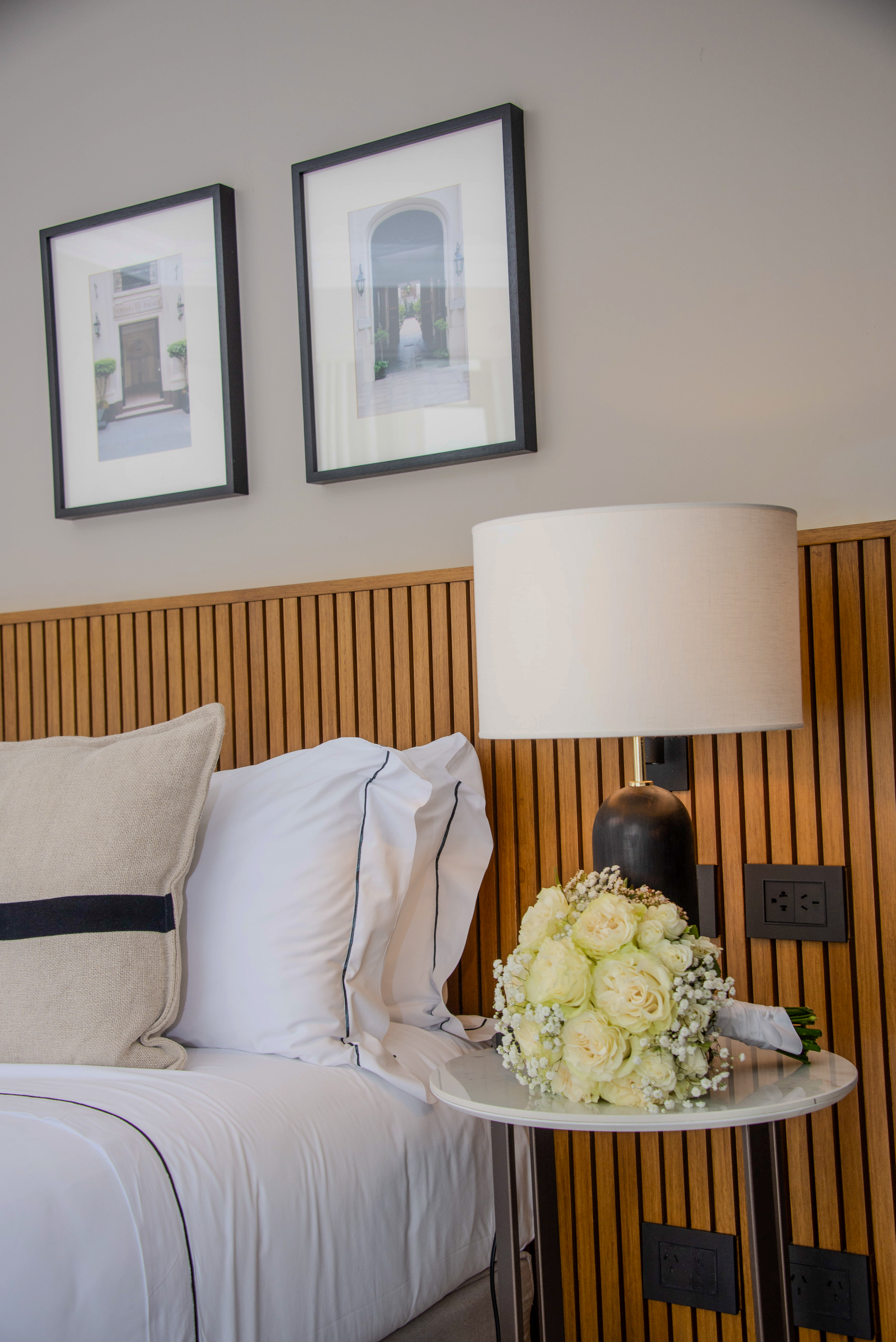 Elegant bedroom with white bed, wooden headboard, lamp, and white rose bouquet.