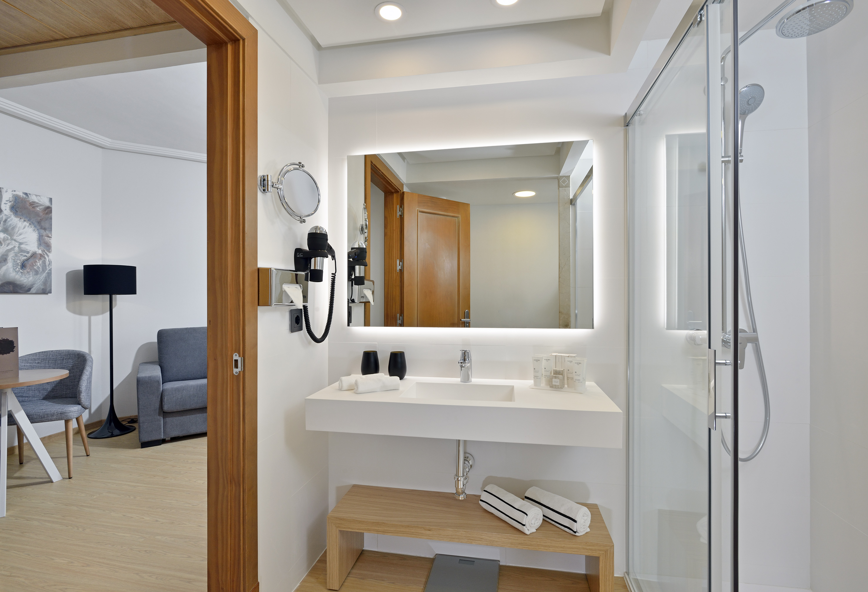 Modern hotel bathroom with lighted mirror, glass shower, and open door to room.