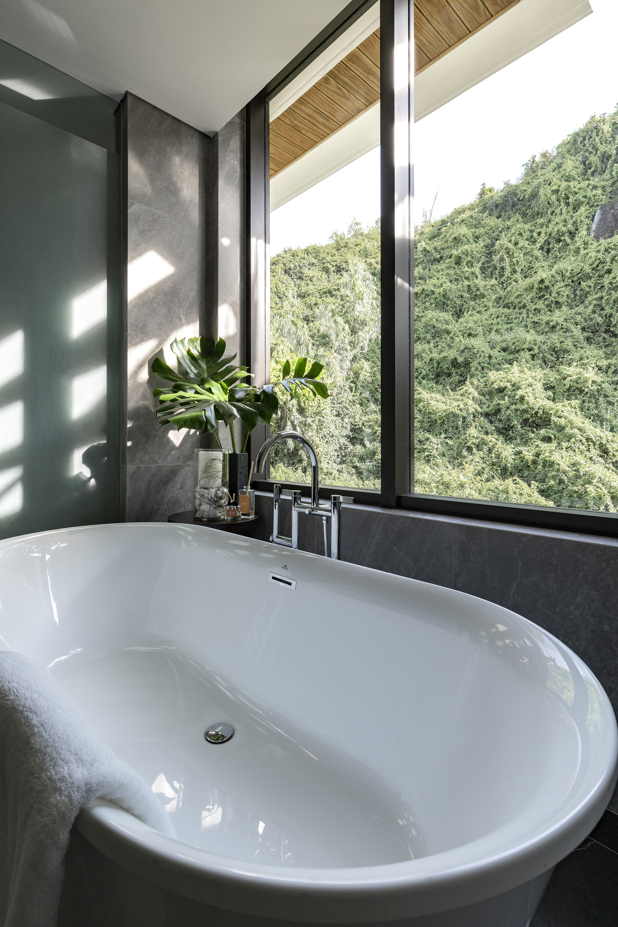 Modern bathroom with white freestanding tub, large window, and green foliage view.