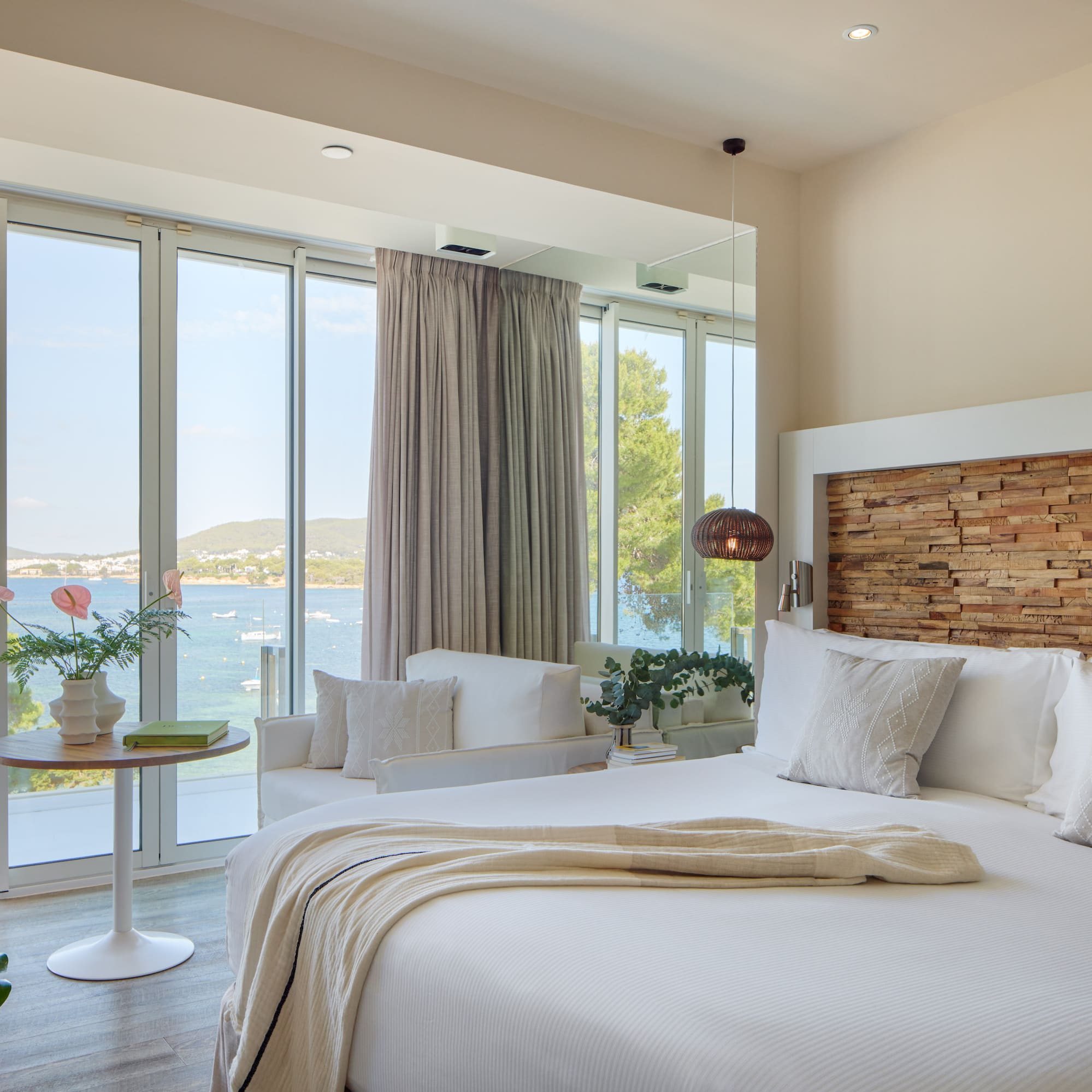 Modern hotel room with sea view, white bed, wooden headboard, panoramic windows.