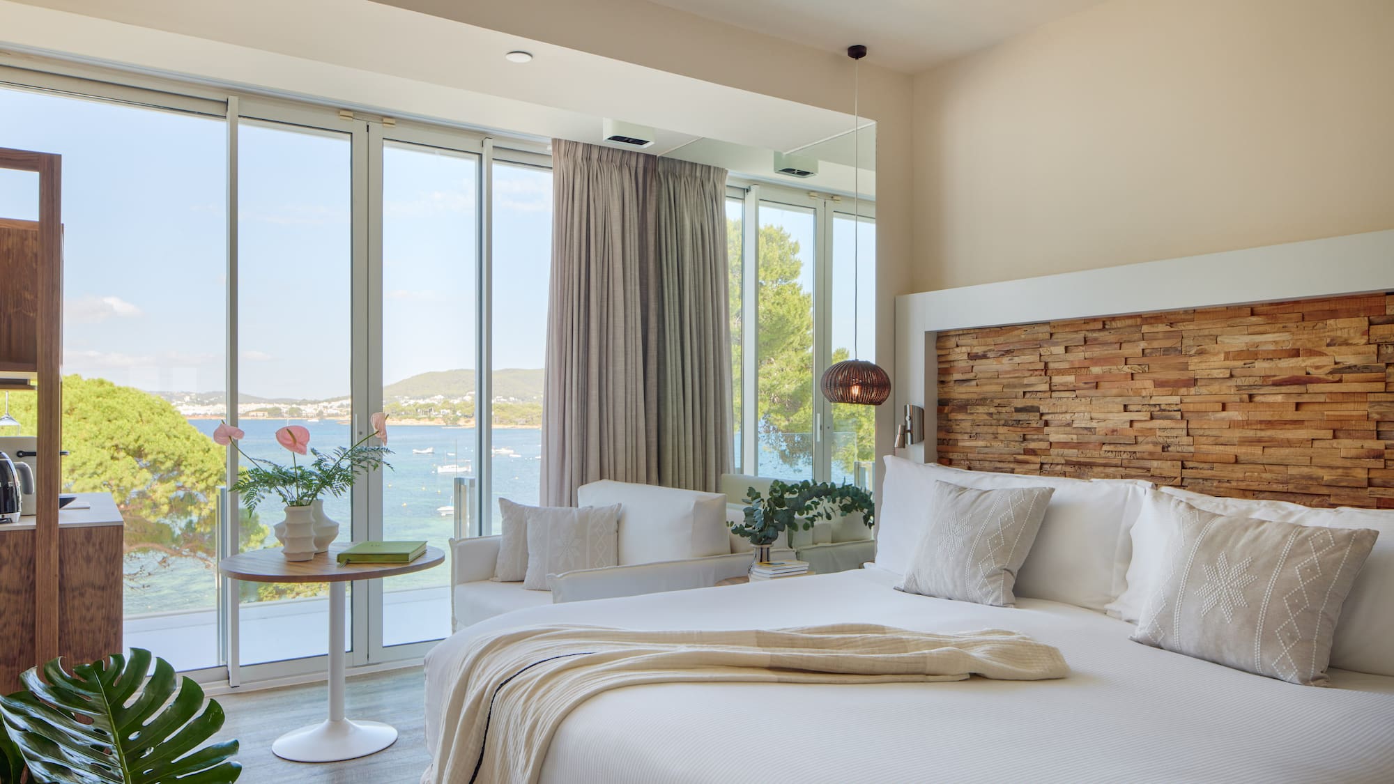 Modern hotel room with sea view, white bed, wooden headboard, panoramic windows.