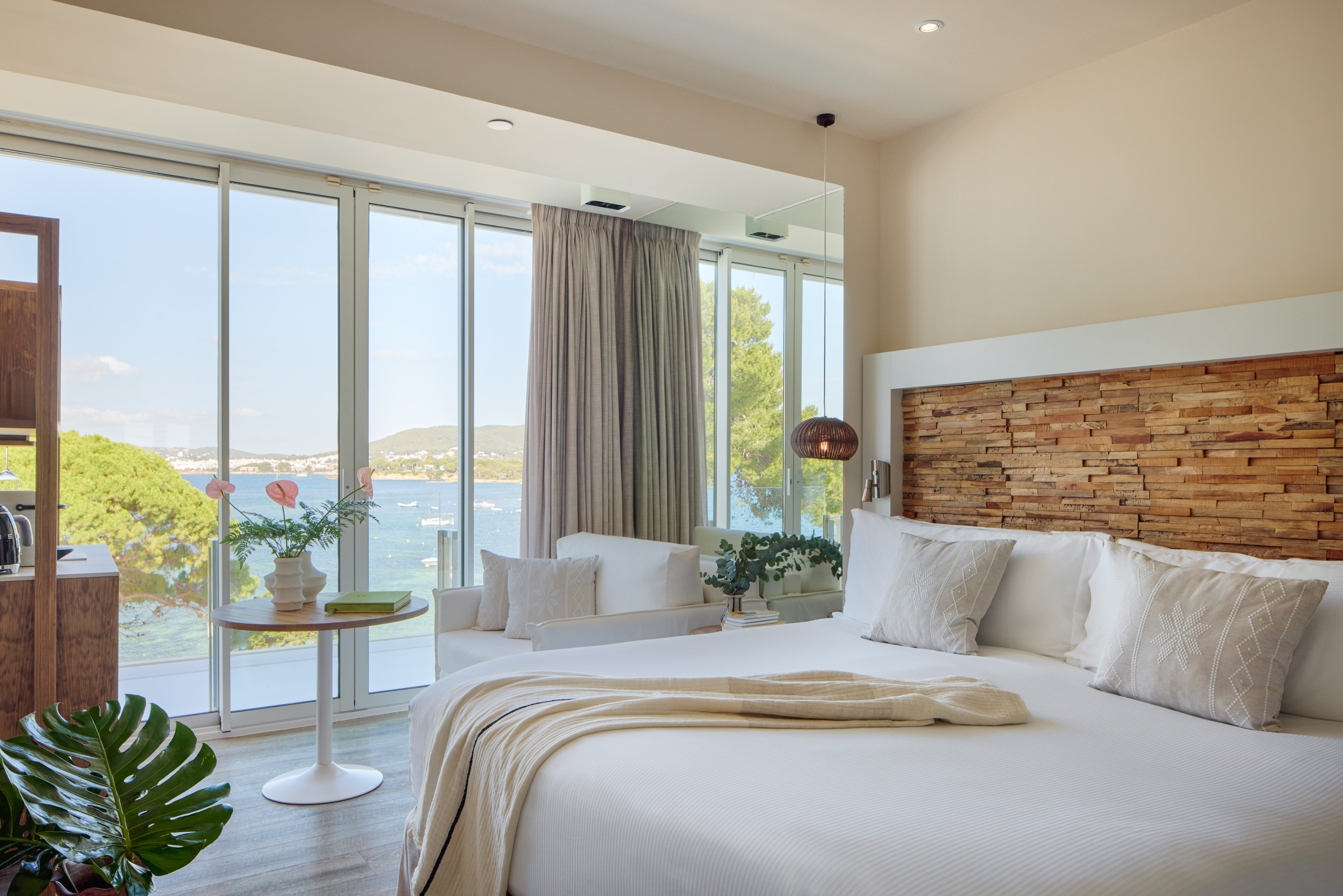 Modern hotel room with sea view, white bed, wooden headboard, panoramic windows.