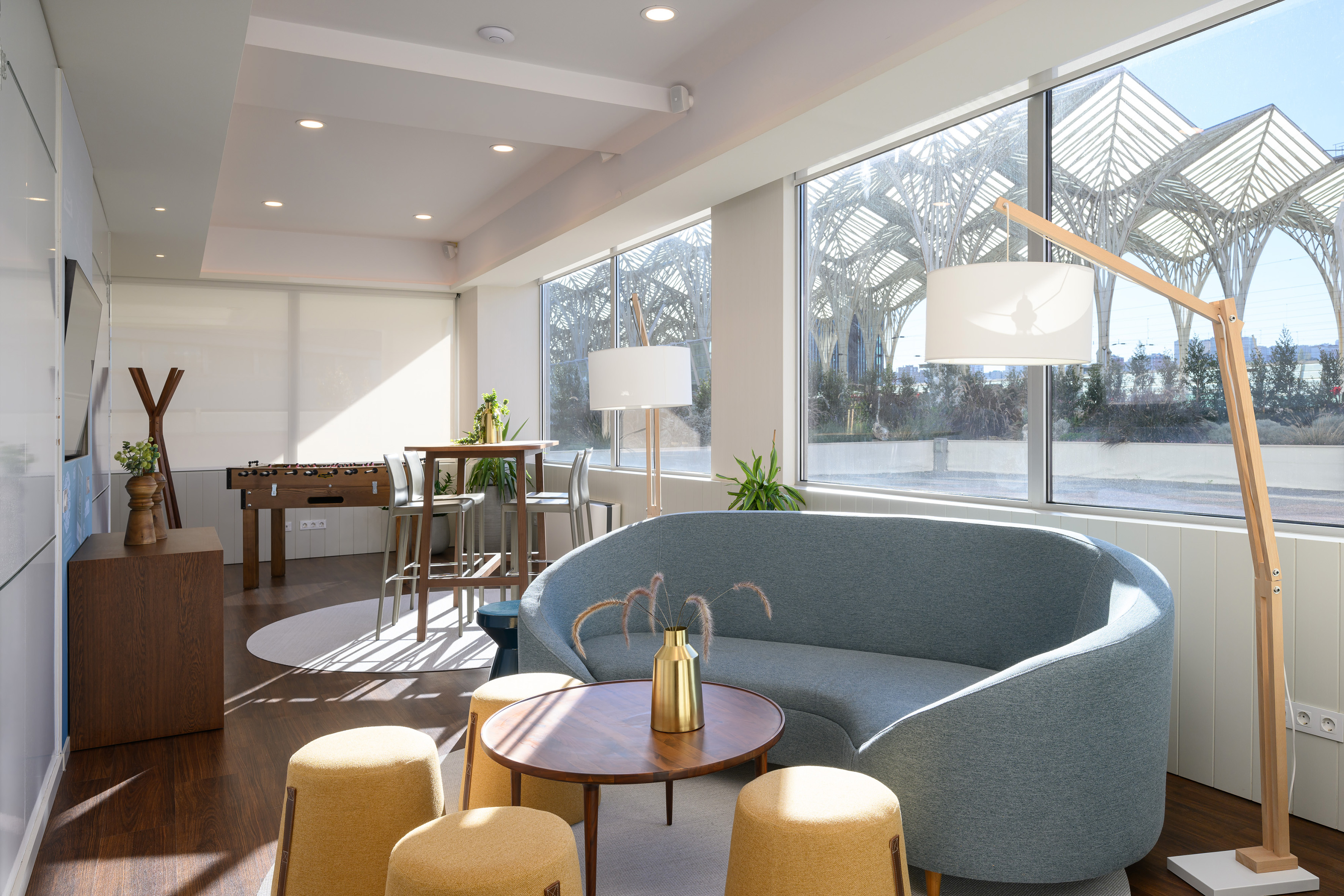 Modern lounge with blue curved sofa, foosball, and large bright windows.