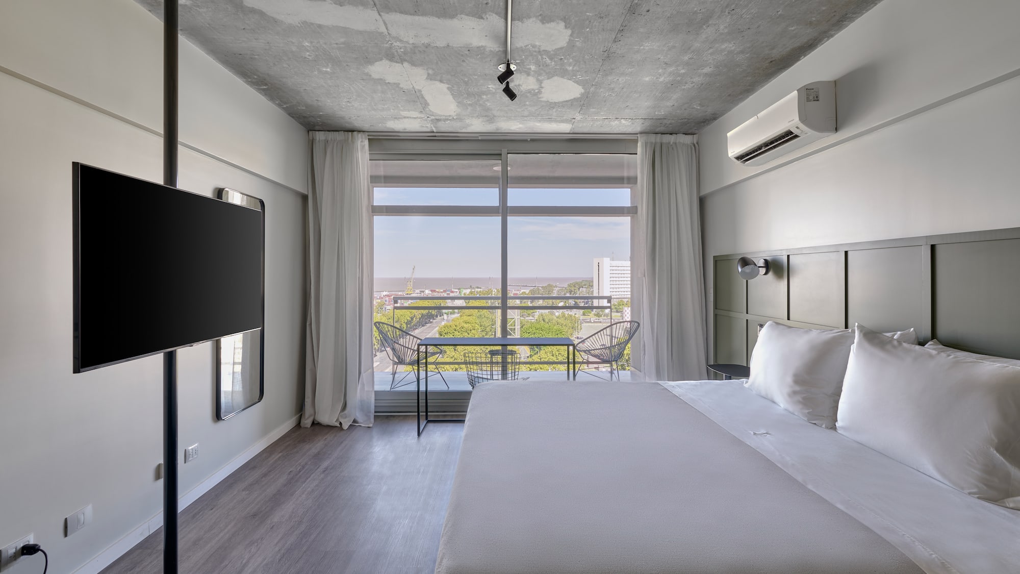 Modern hotel room with concrete ceiling, large bed, balcony, and city view.