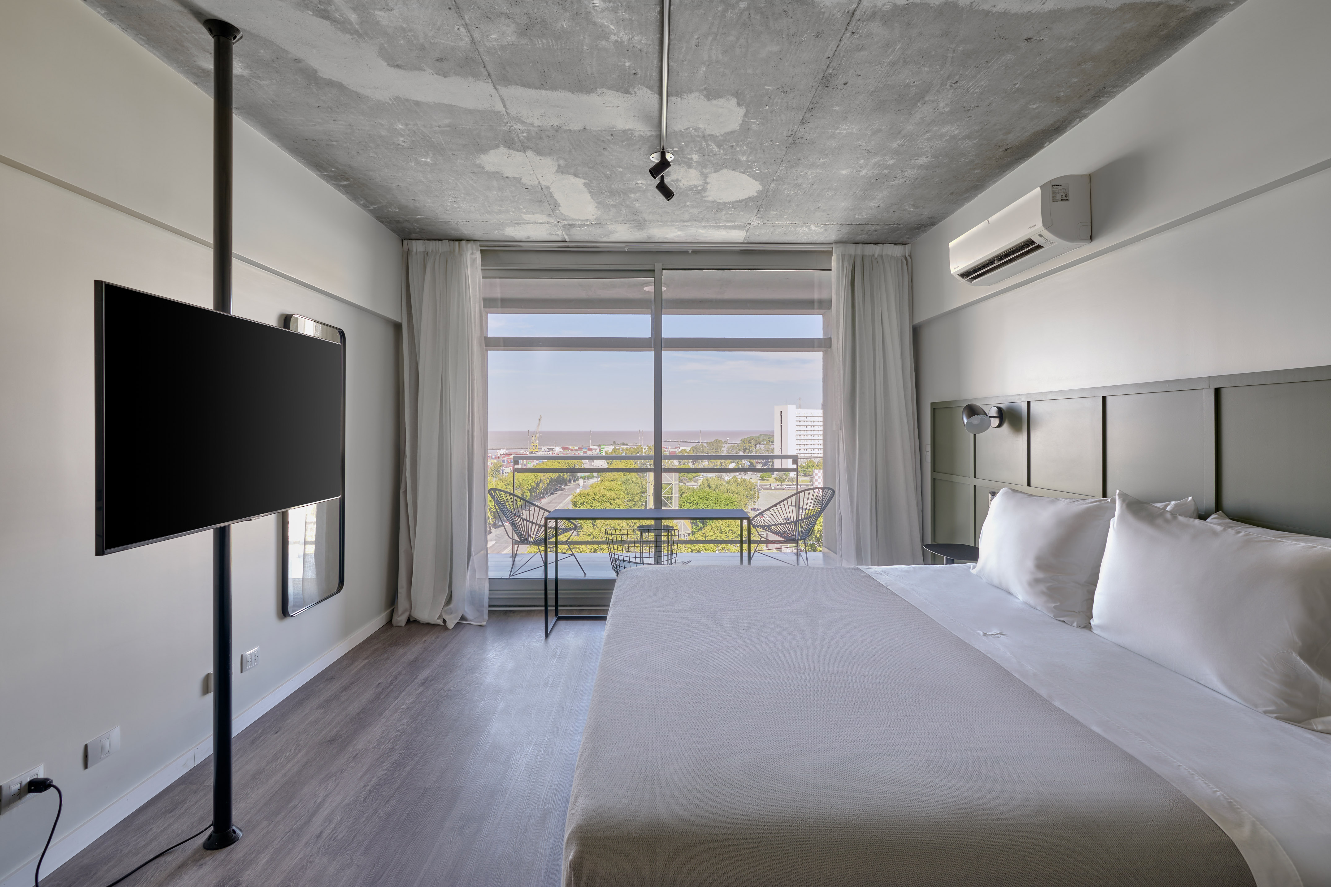 Modern hotel room with concrete ceiling, large bed, balcony, and city view.