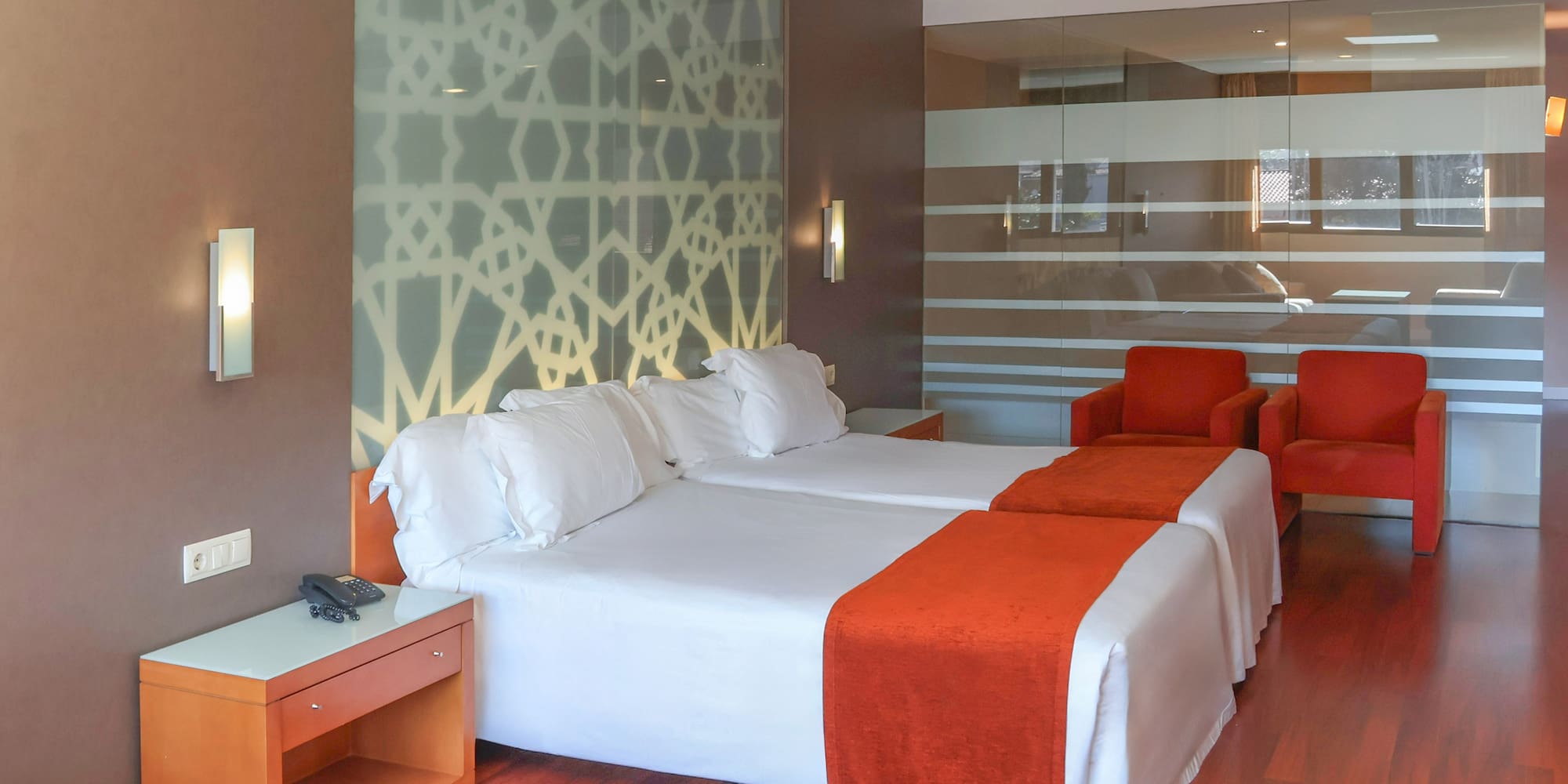 Modern hotel room with twin beds, orange runners, geometric wall, red chairs.