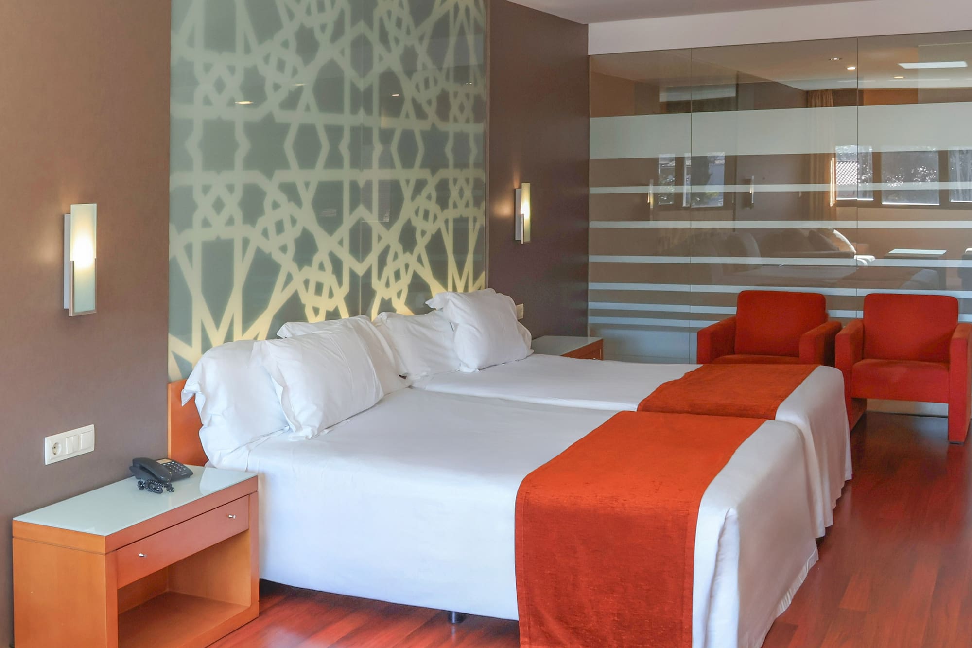 Modern hotel room with twin beds, orange runners, geometric wall, red chairs.