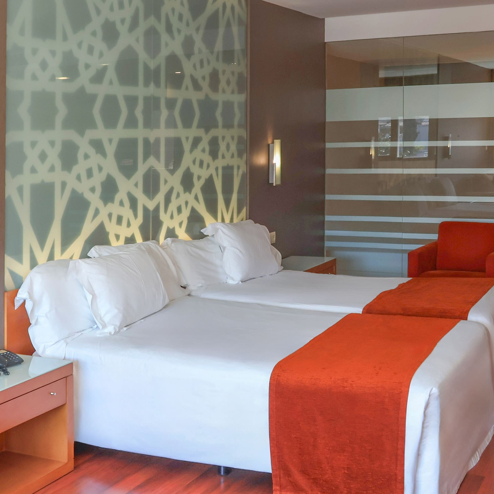 Modern hotel room with twin beds, orange runners, geometric wall, red chairs.