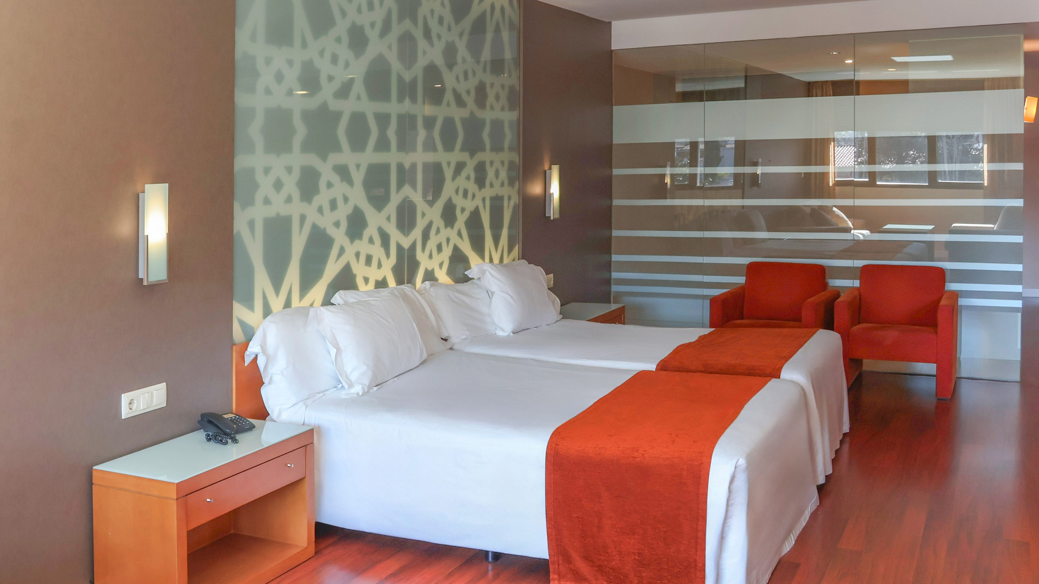 Modern hotel room with twin beds, orange runners, geometric wall, red chairs.