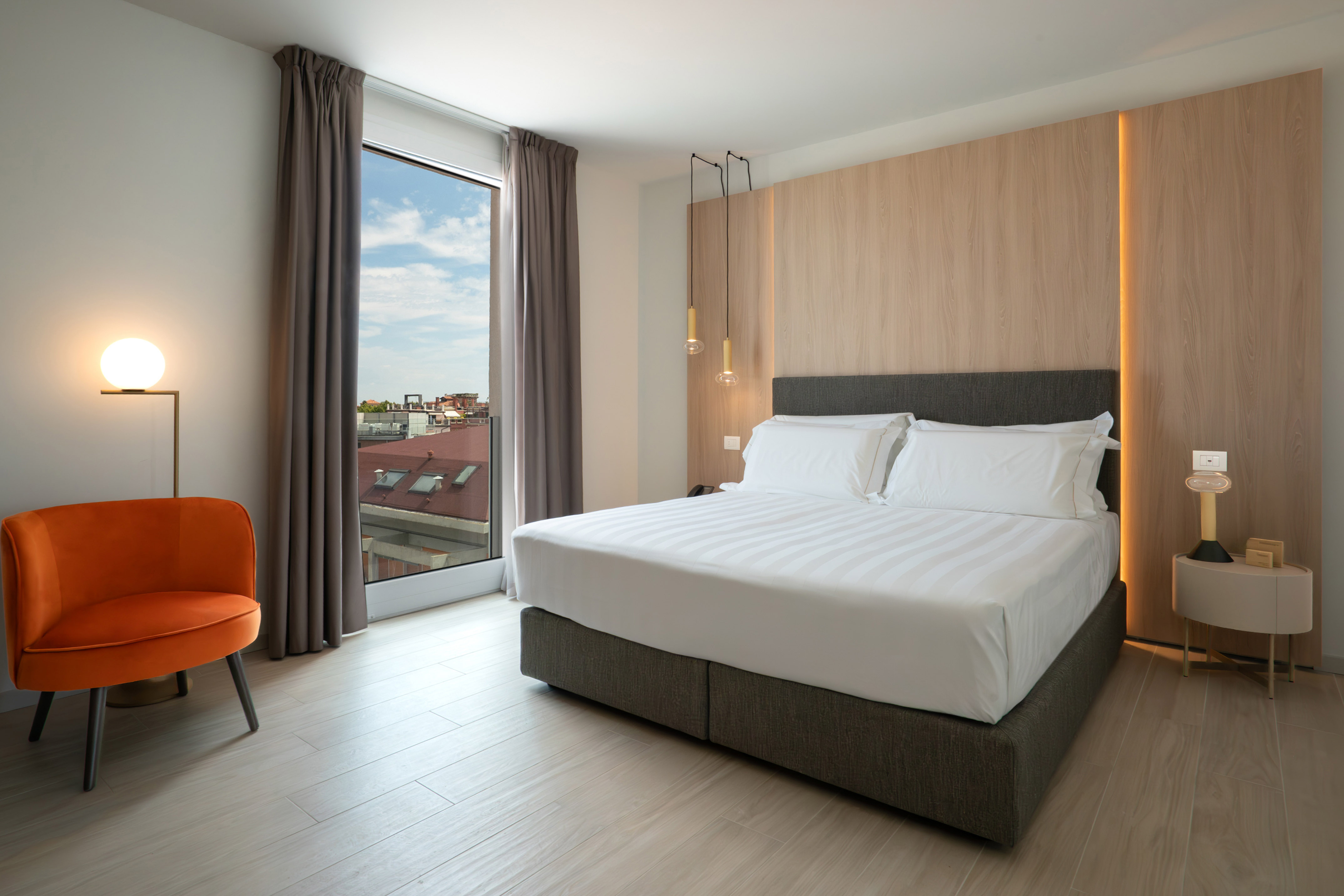 Modern hotel bedroom with queen bed, orange armchair, wood accent wall, city view.