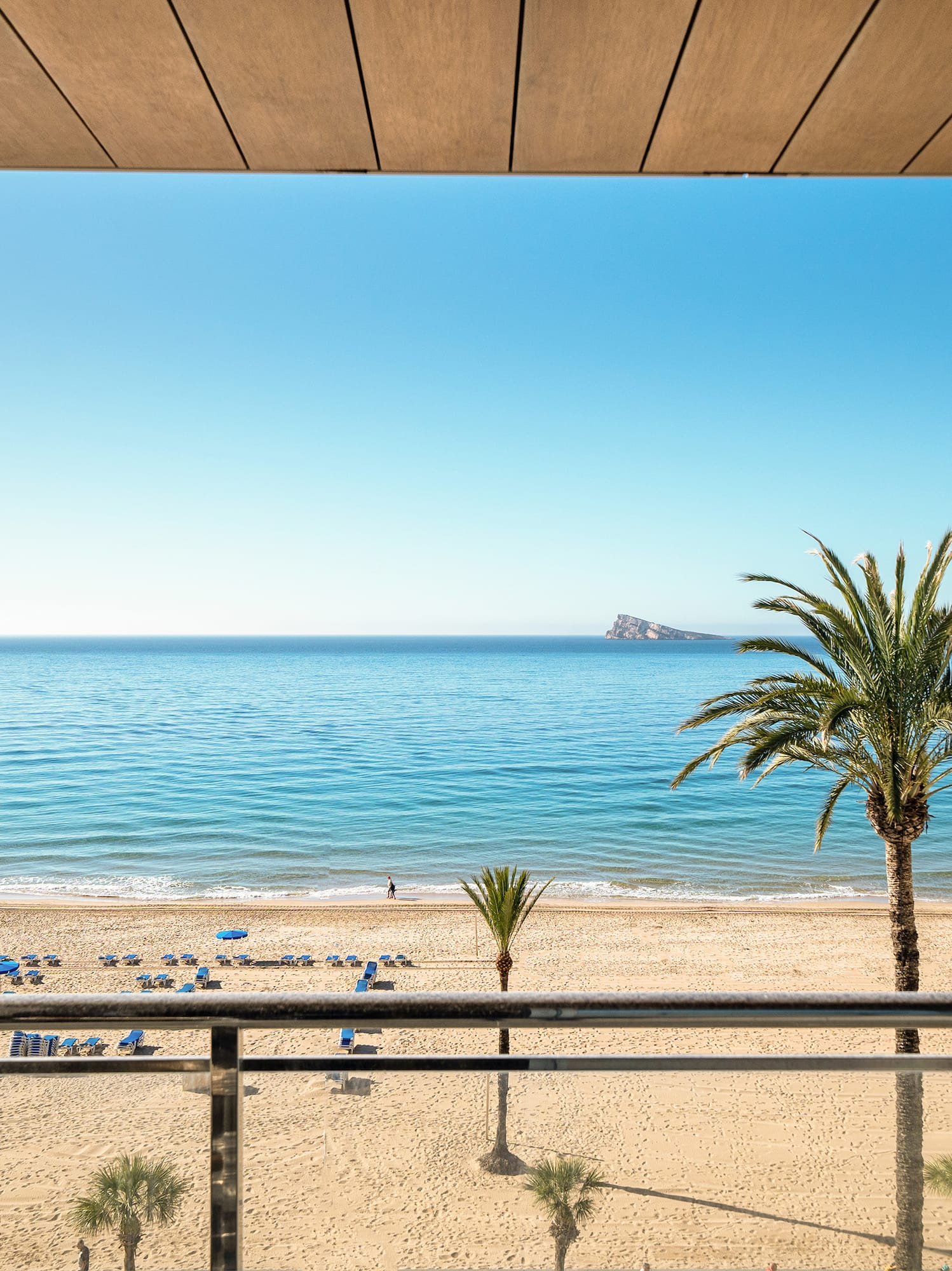 Benidorm beach from hotel balcony: sunny sea, sand, palm trees, distant island.