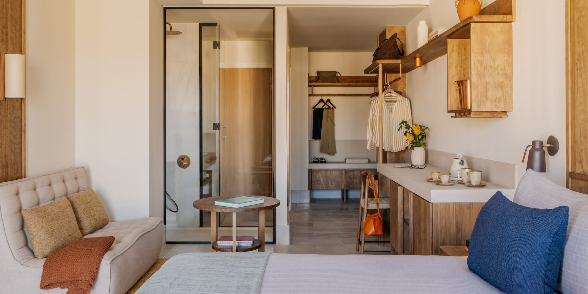 Modern hotel room with bed, glass shower, open closet, and wood details.
