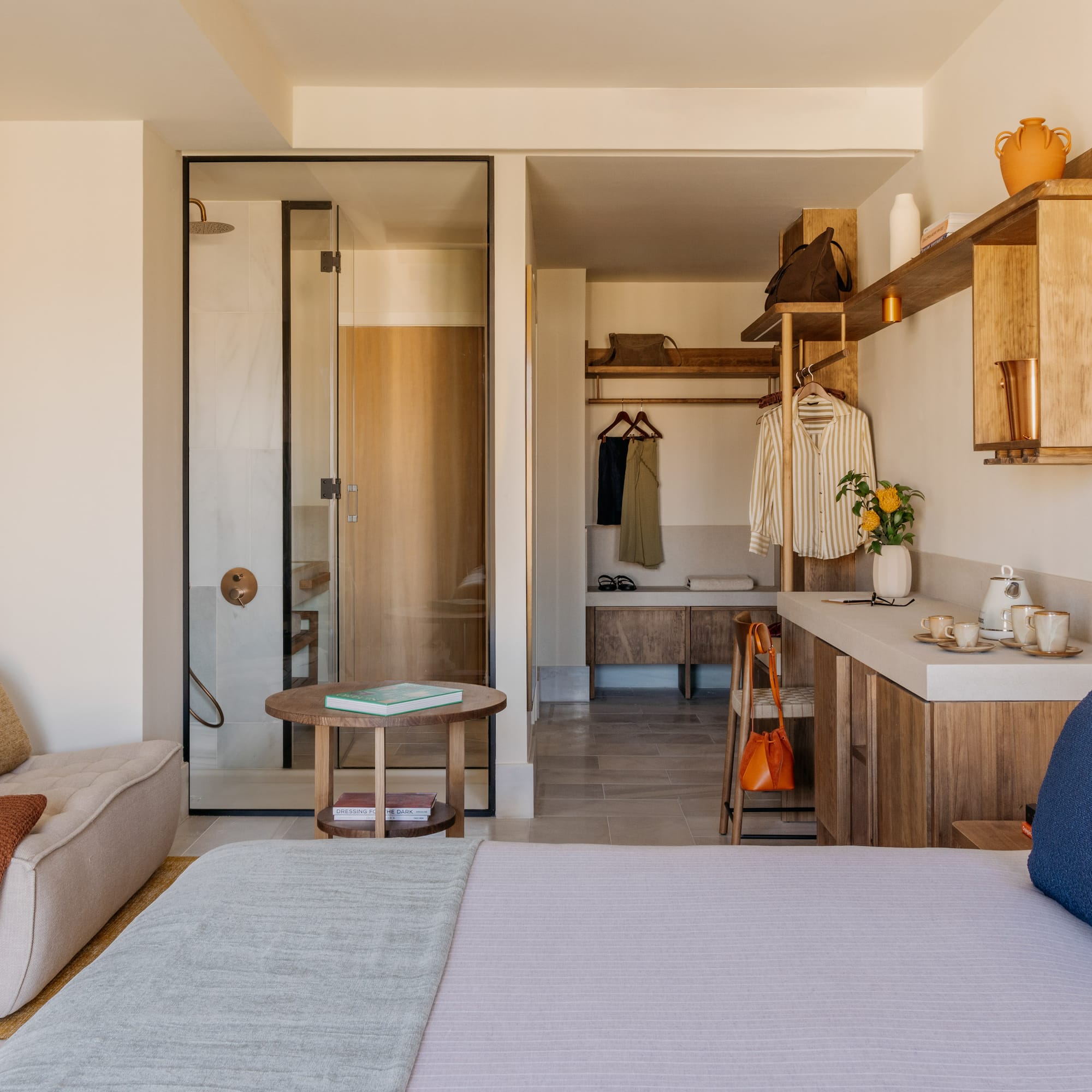 Modern hotel room with bed, glass shower, open closet, and wood details.