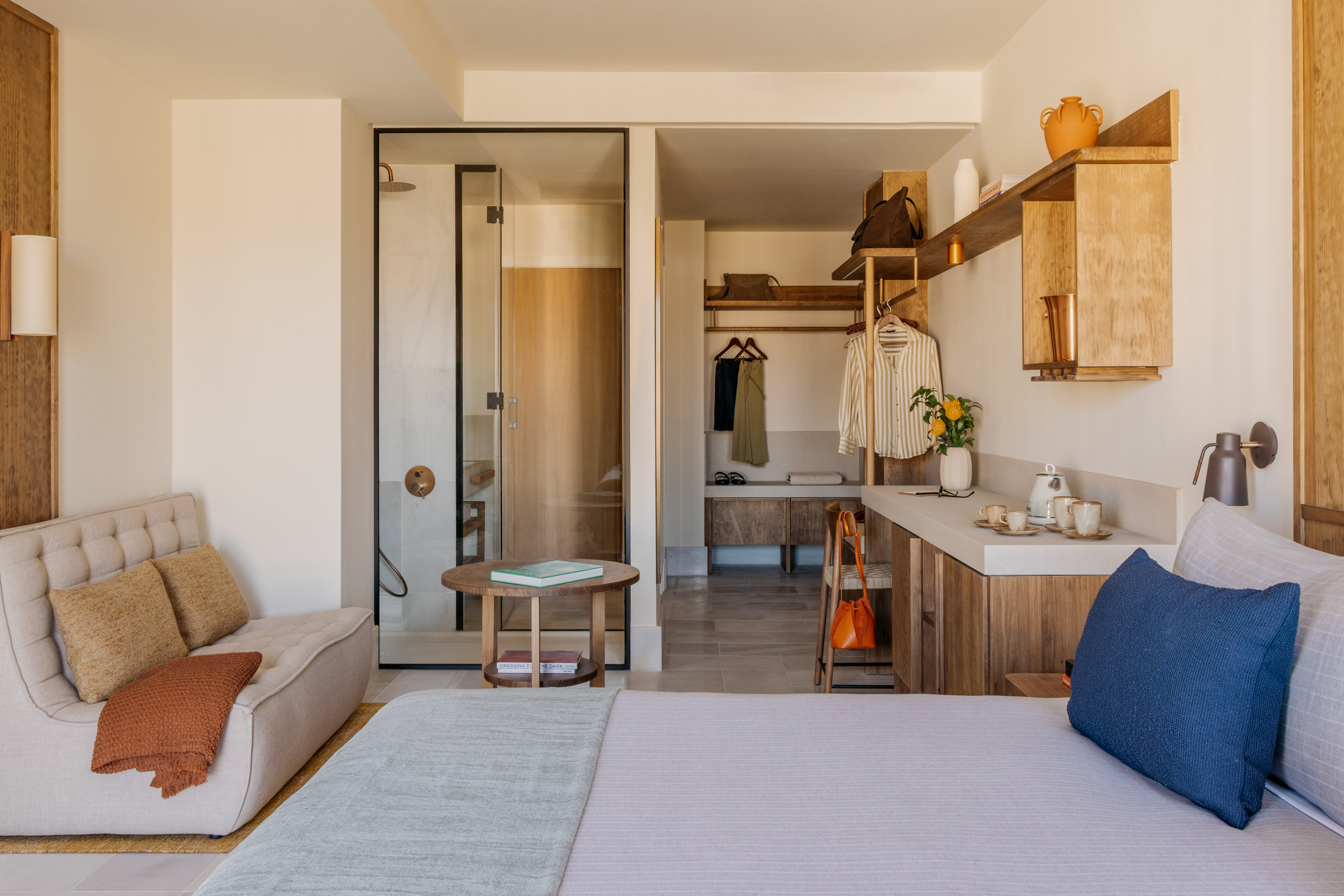 Modern hotel room with bed, glass shower, open closet, and wood details.