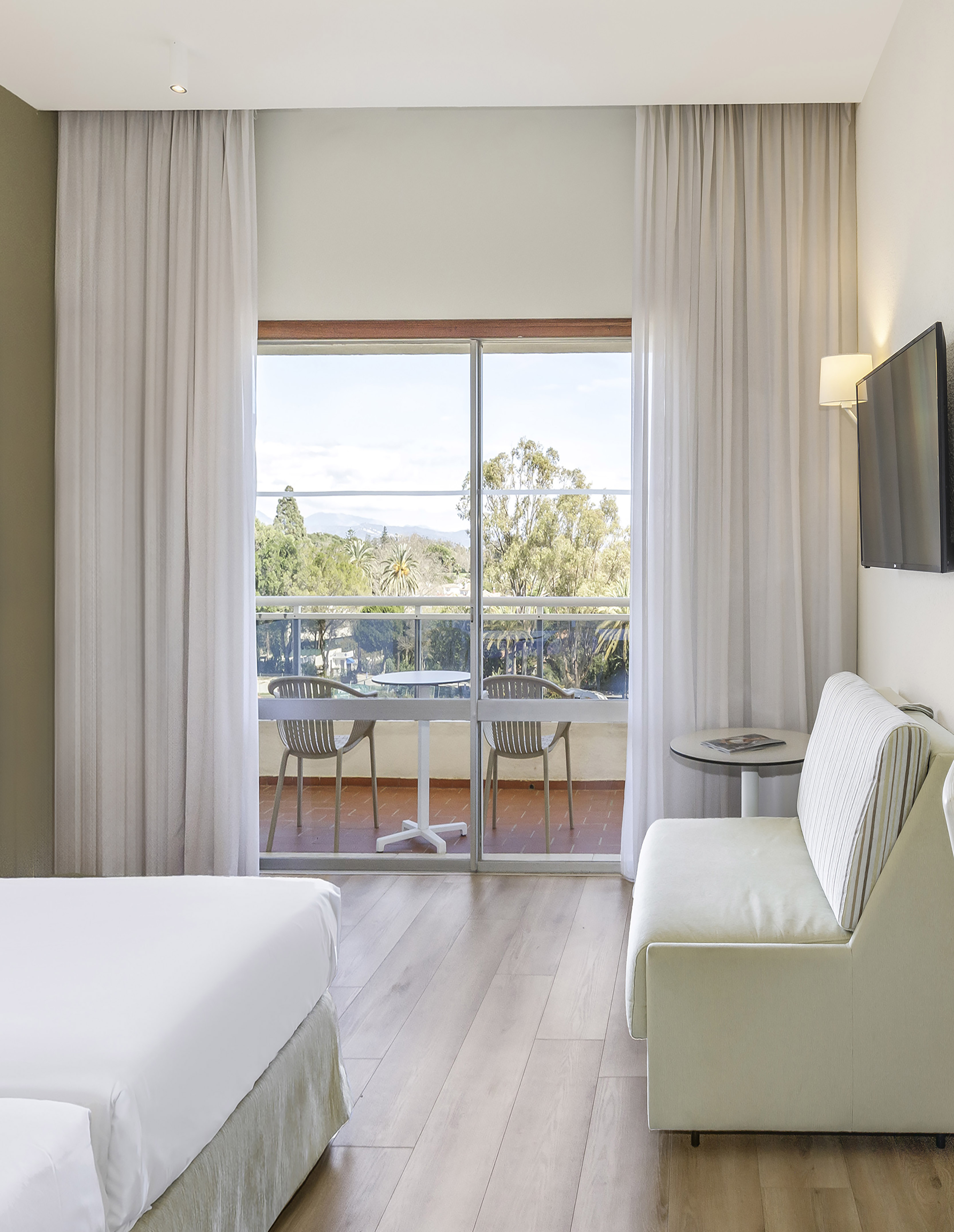 Bright hotel room with balcony, green landscape view, white bed, and modern sofa.