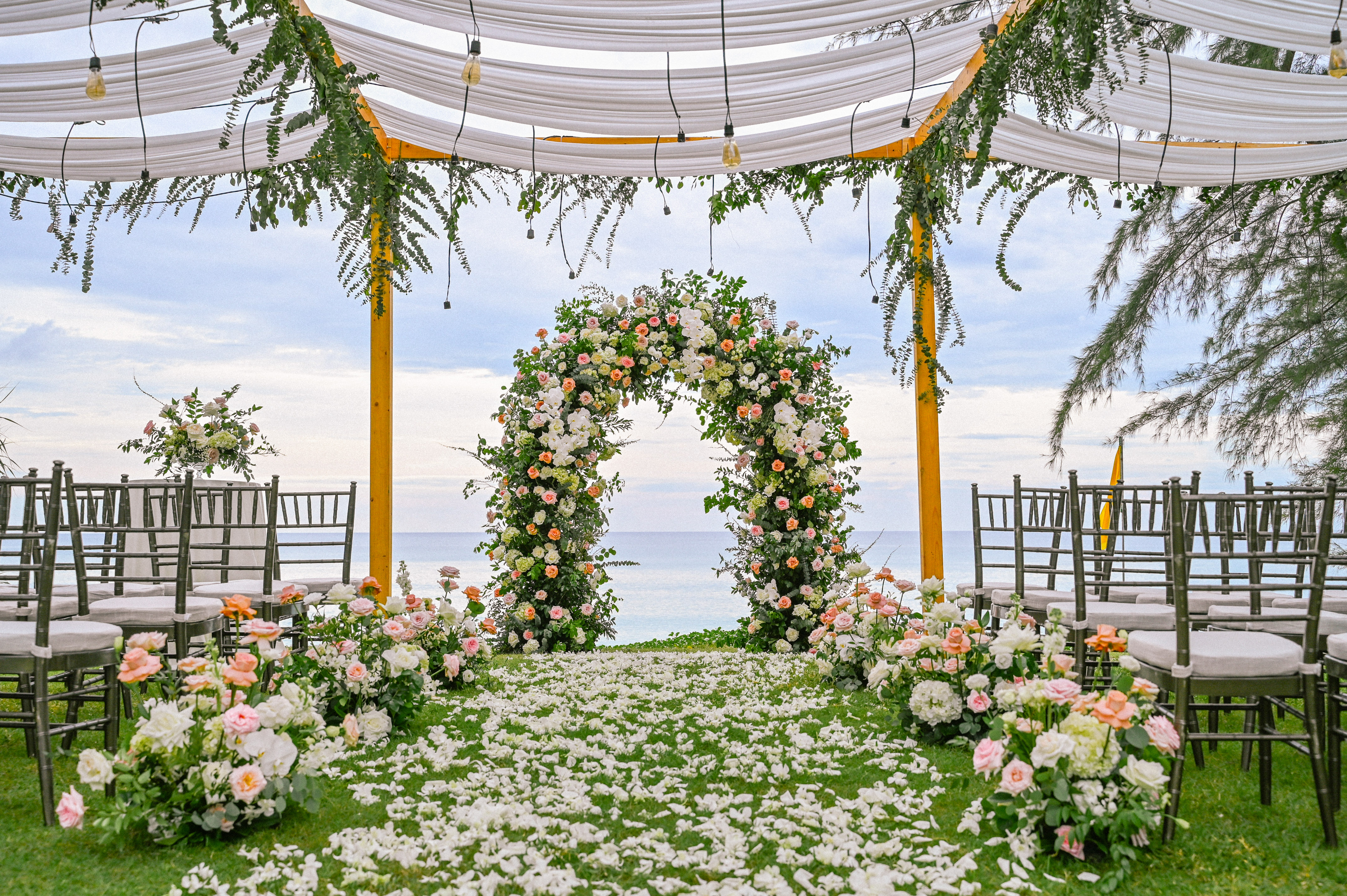 Beach wedding ceremony setup with floral arch, rose petals aisle, and ocean view.