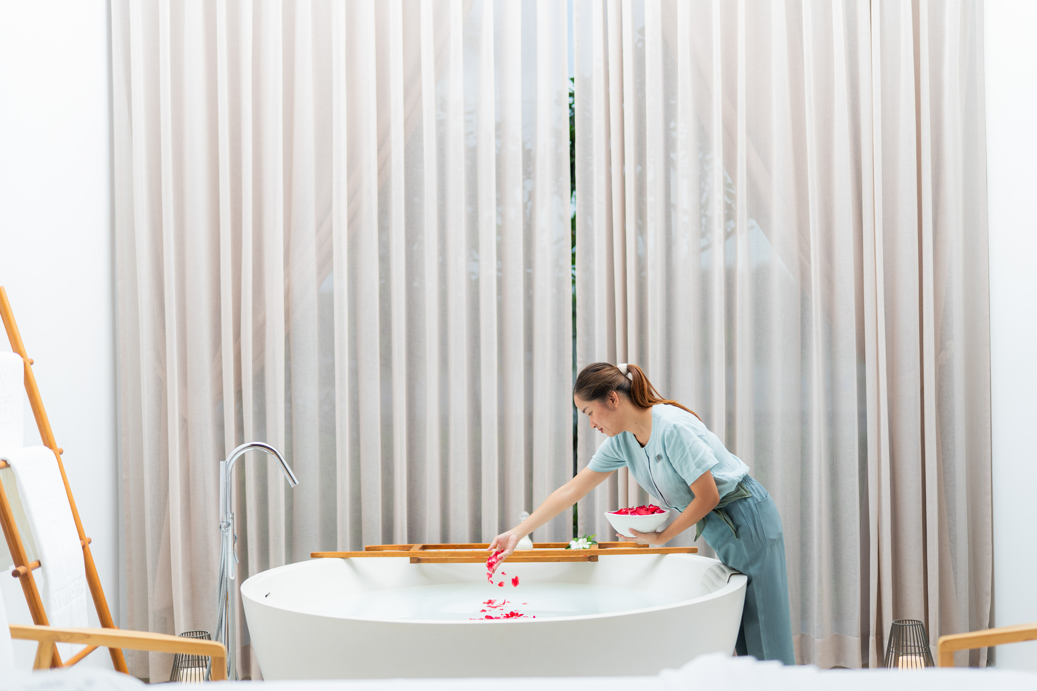 Woman preparing luxury spa bath, adding red rose petals to white tub.