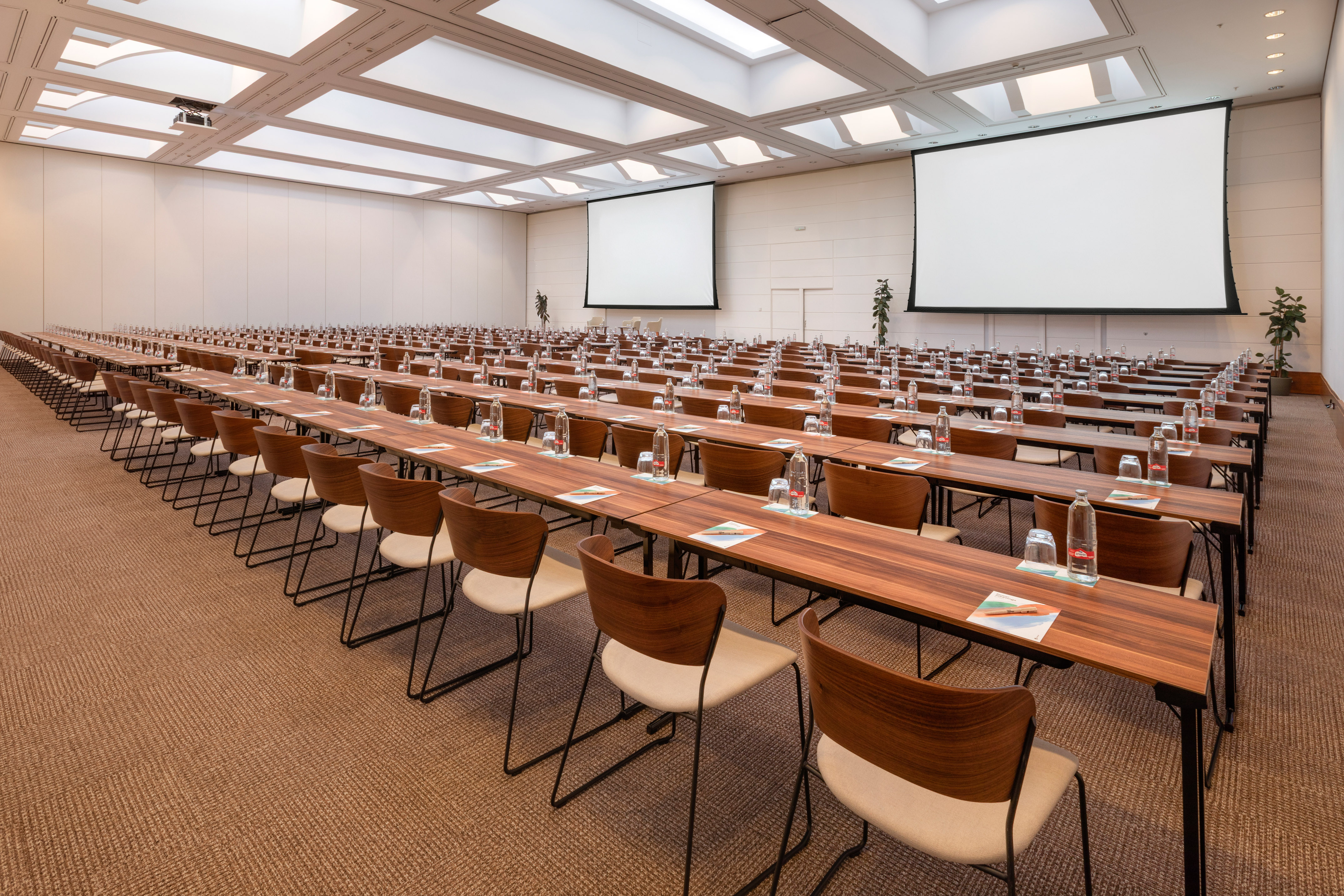 Empty conference room with rows of tables, chairs, water bottles, and two projector screens.