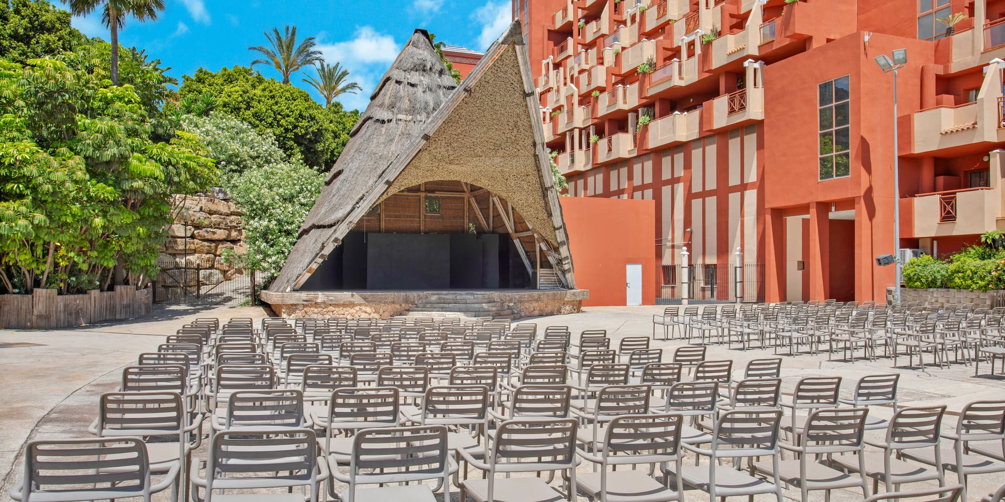 Outdoor amphitheater with thatched stage and rows of empty chairs at a vibrant resort.