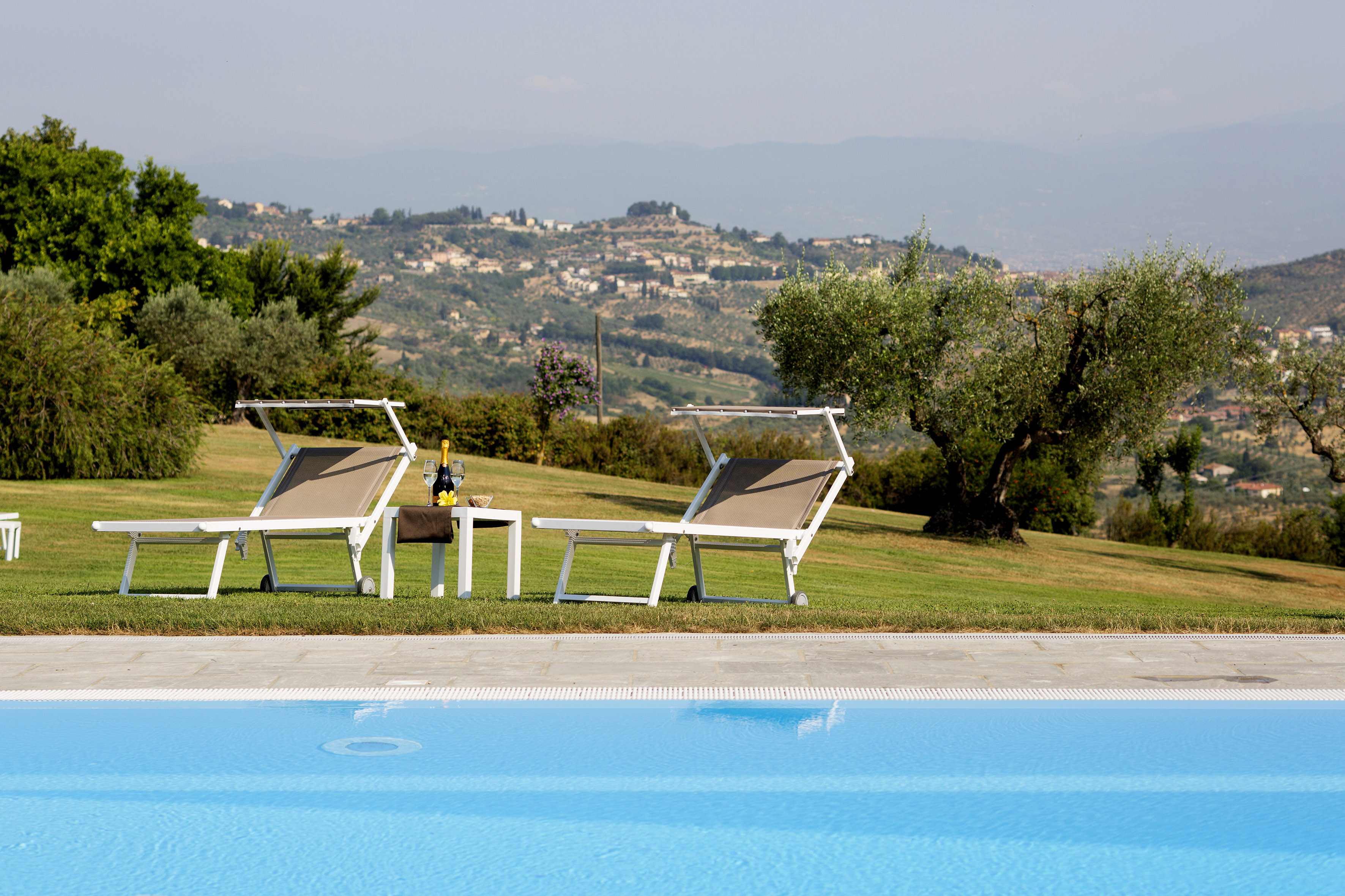 Poolside sun loungers, champagne, and scenic Tuscan hills with villages.