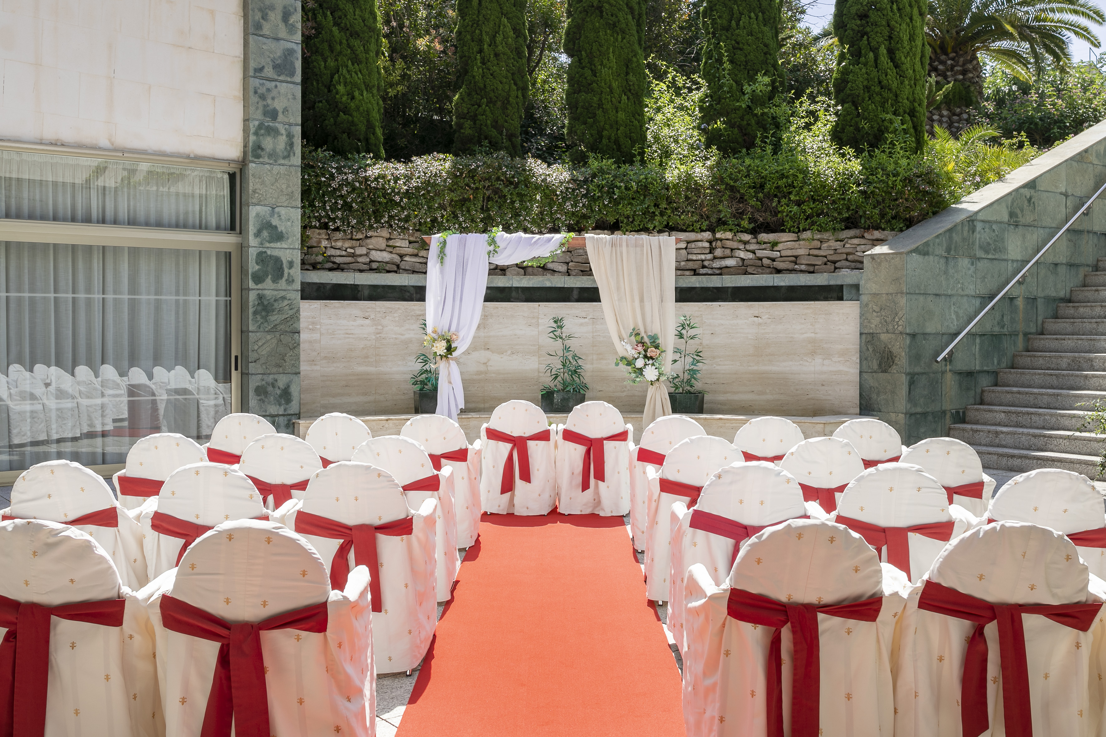 Outdoor wedding ceremony aisle with red carpet, white chairs, floral backdrop.