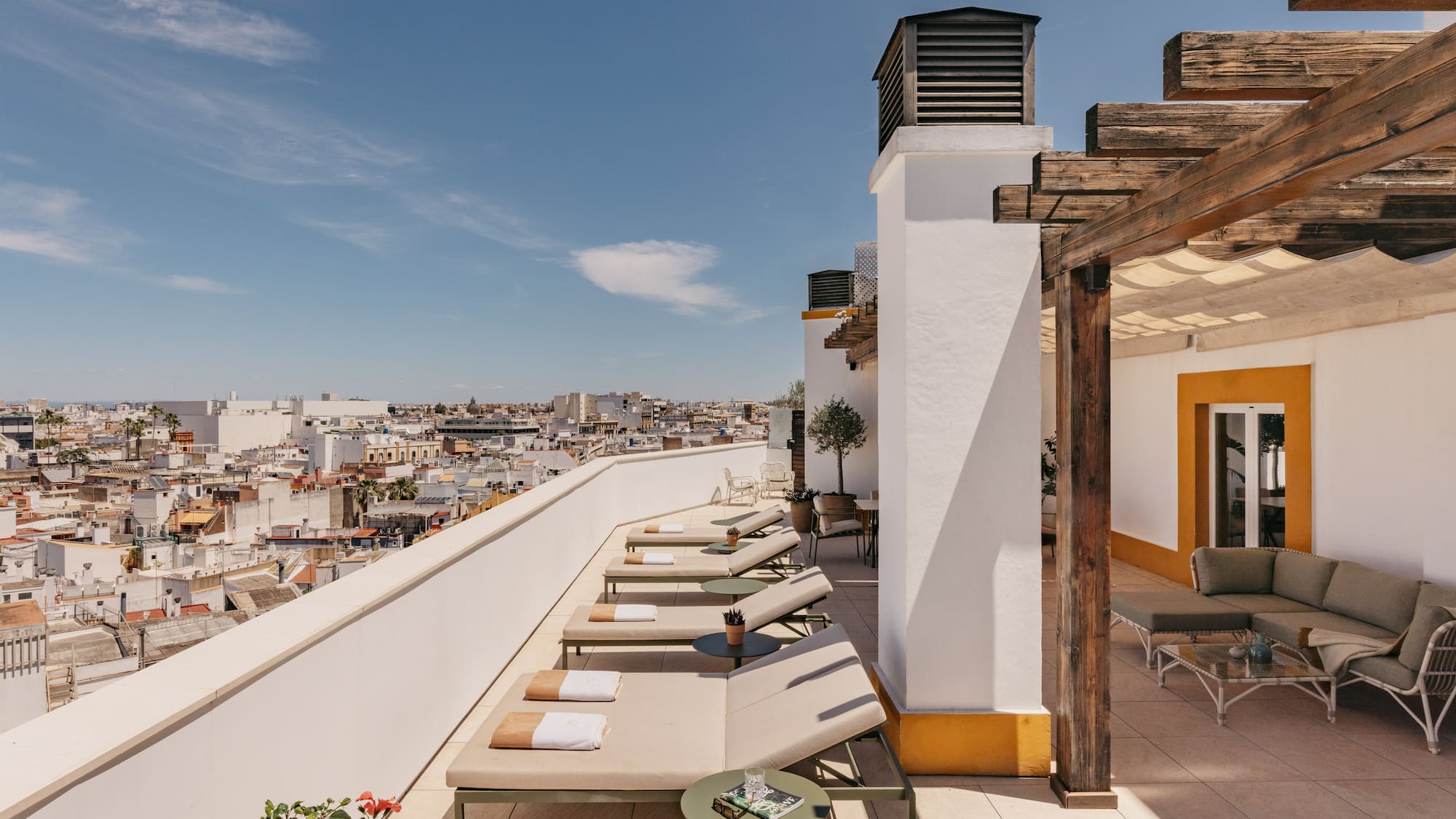 Azotea mediterránea con tumbonas, pérgola de madera y vistas urbanas soleadas.