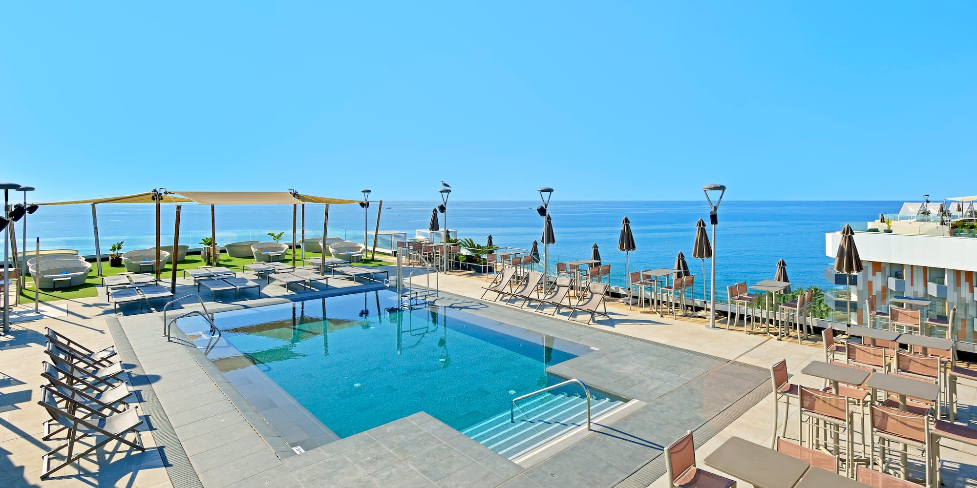 Sunny rooftop pool with ocean view, lounge chairs, and outdoor dining tables.
