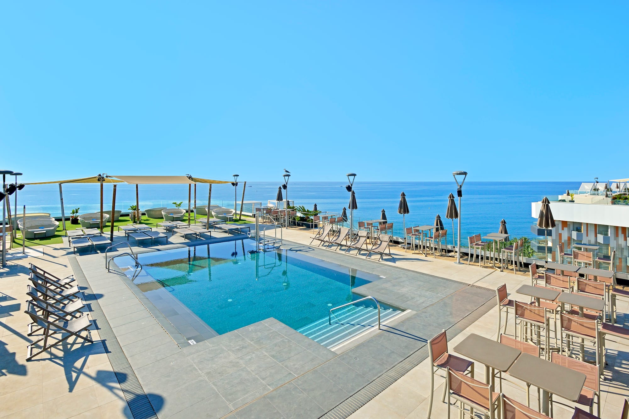 Hotel rooftop pool with ocean view, sunbeds, and outdoor tables.