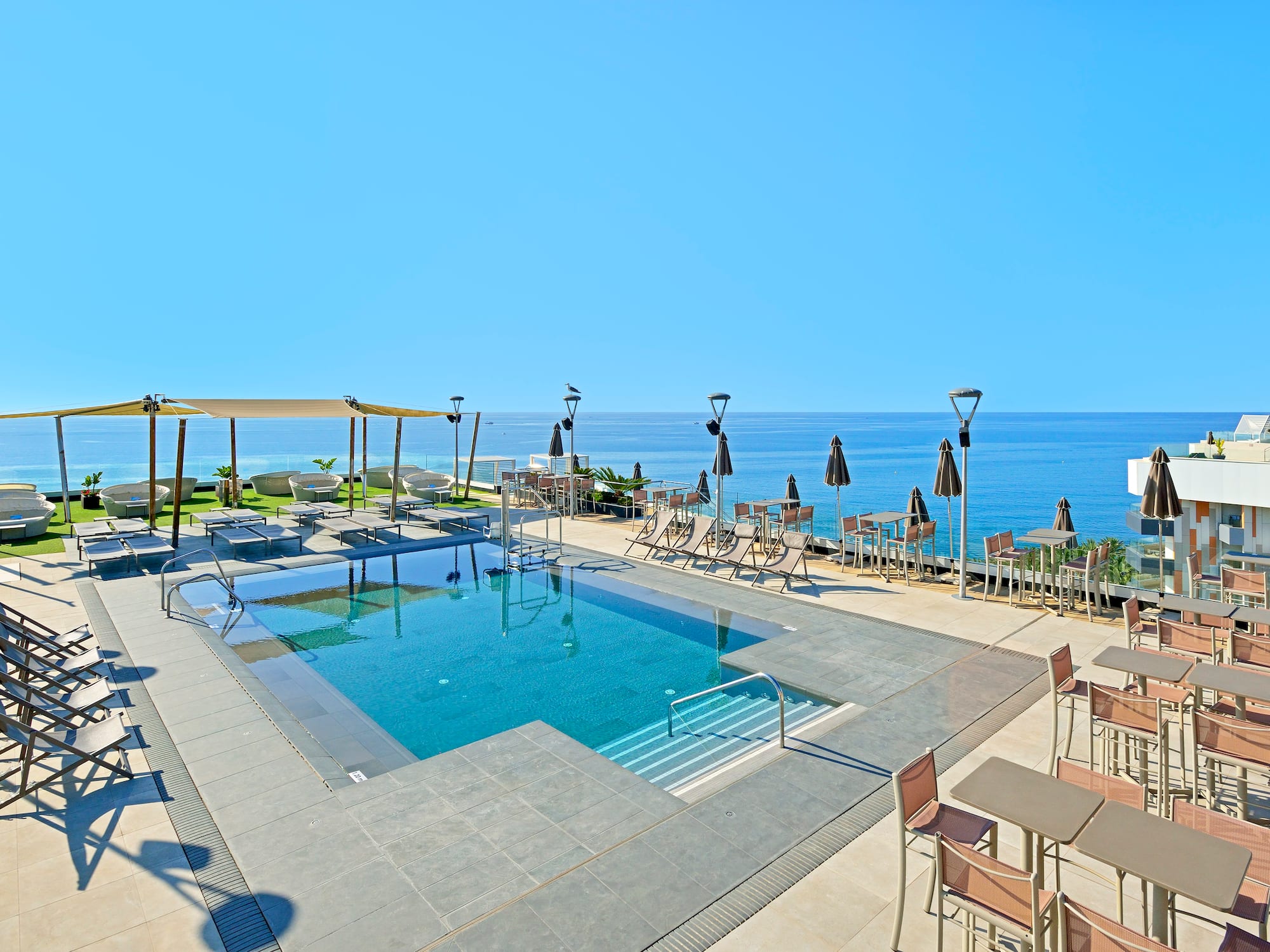 Hotel rooftop pool with ocean view, sunbeds, and outdoor tables.