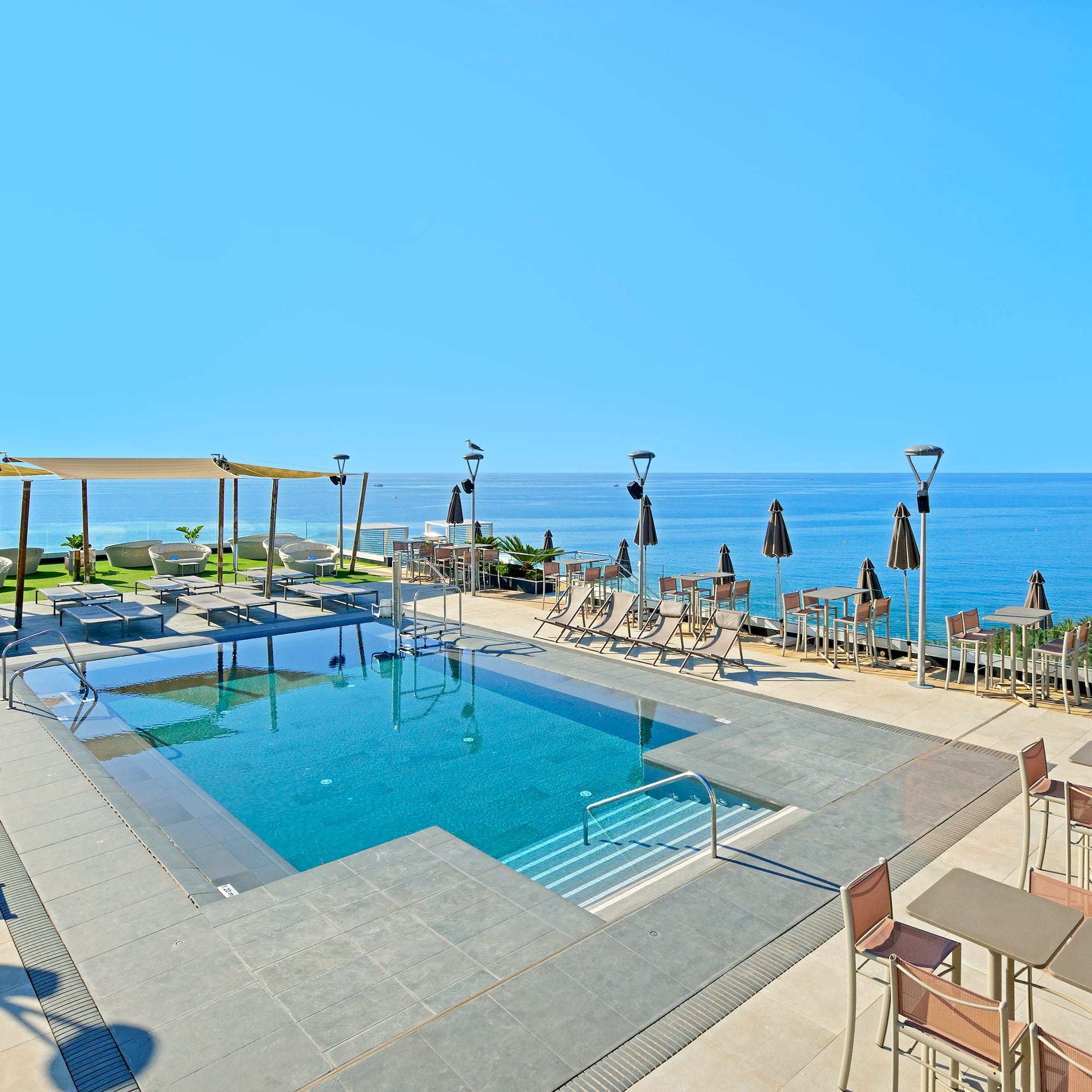 Sunny rooftop pool with ocean view, lounge chairs, and outdoor dining tables.