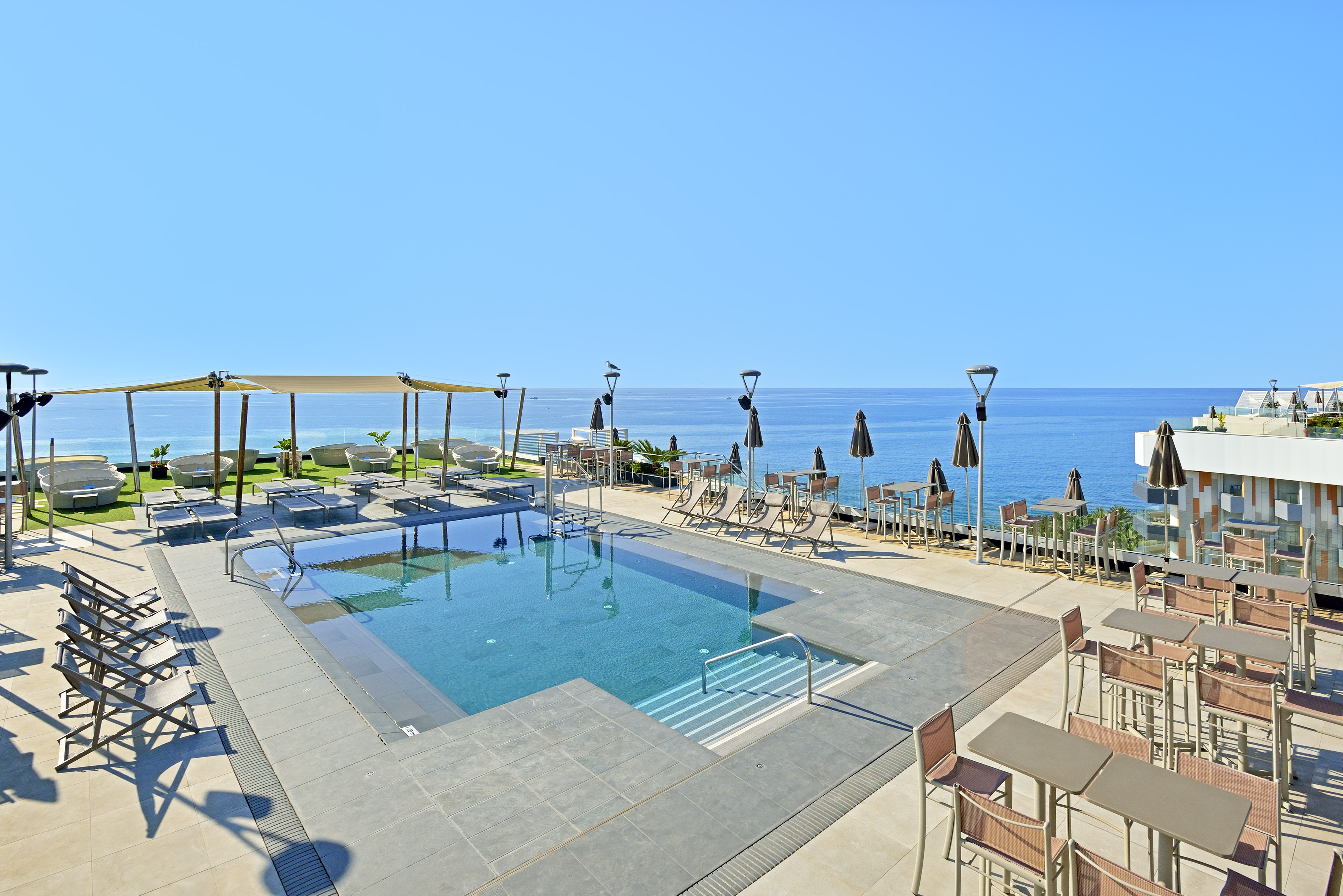 Hotel rooftop pool with ocean view, sunbeds, and outdoor tables.