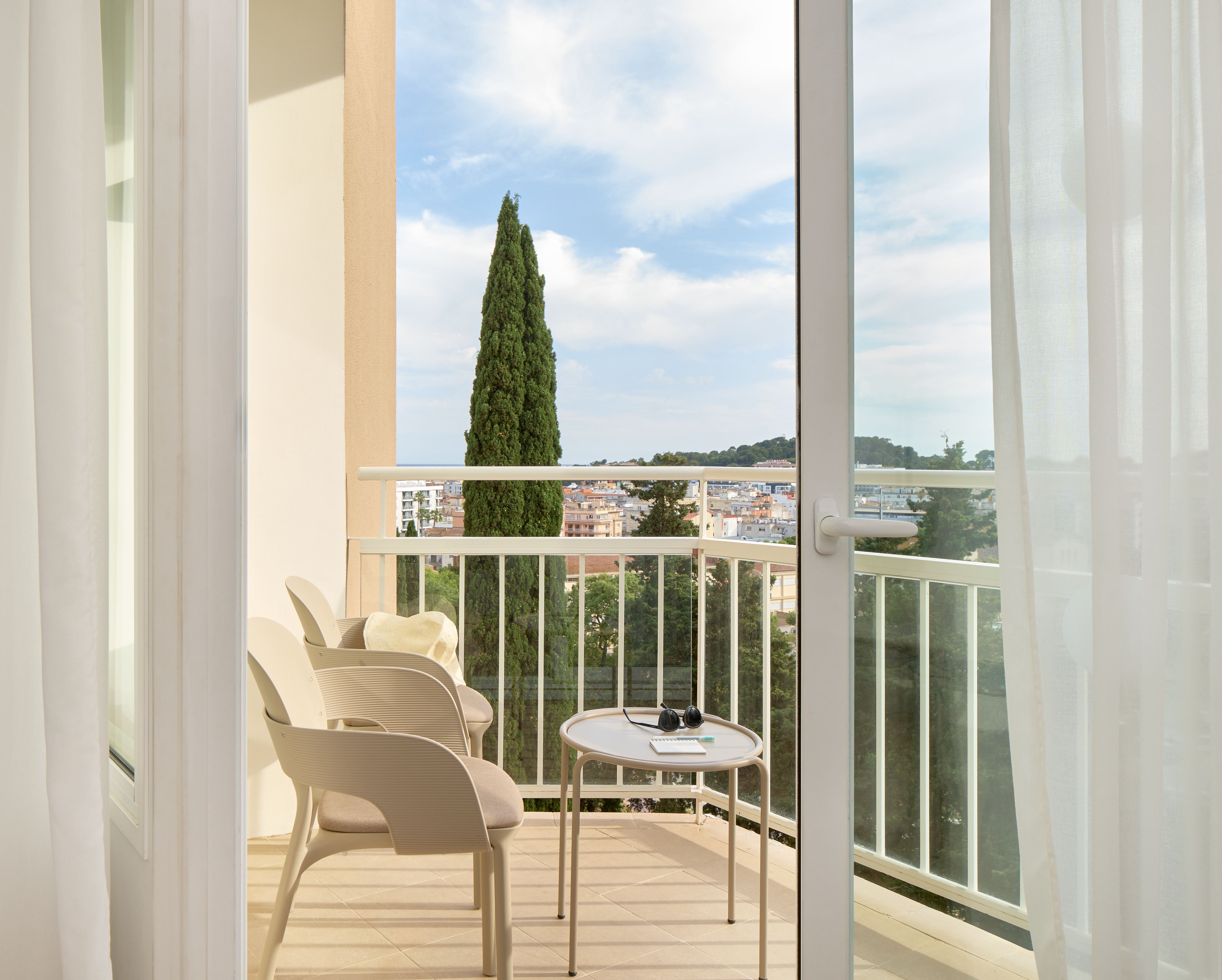 Sunny balcony with outdoor seating, town view, and tall cypress tree.