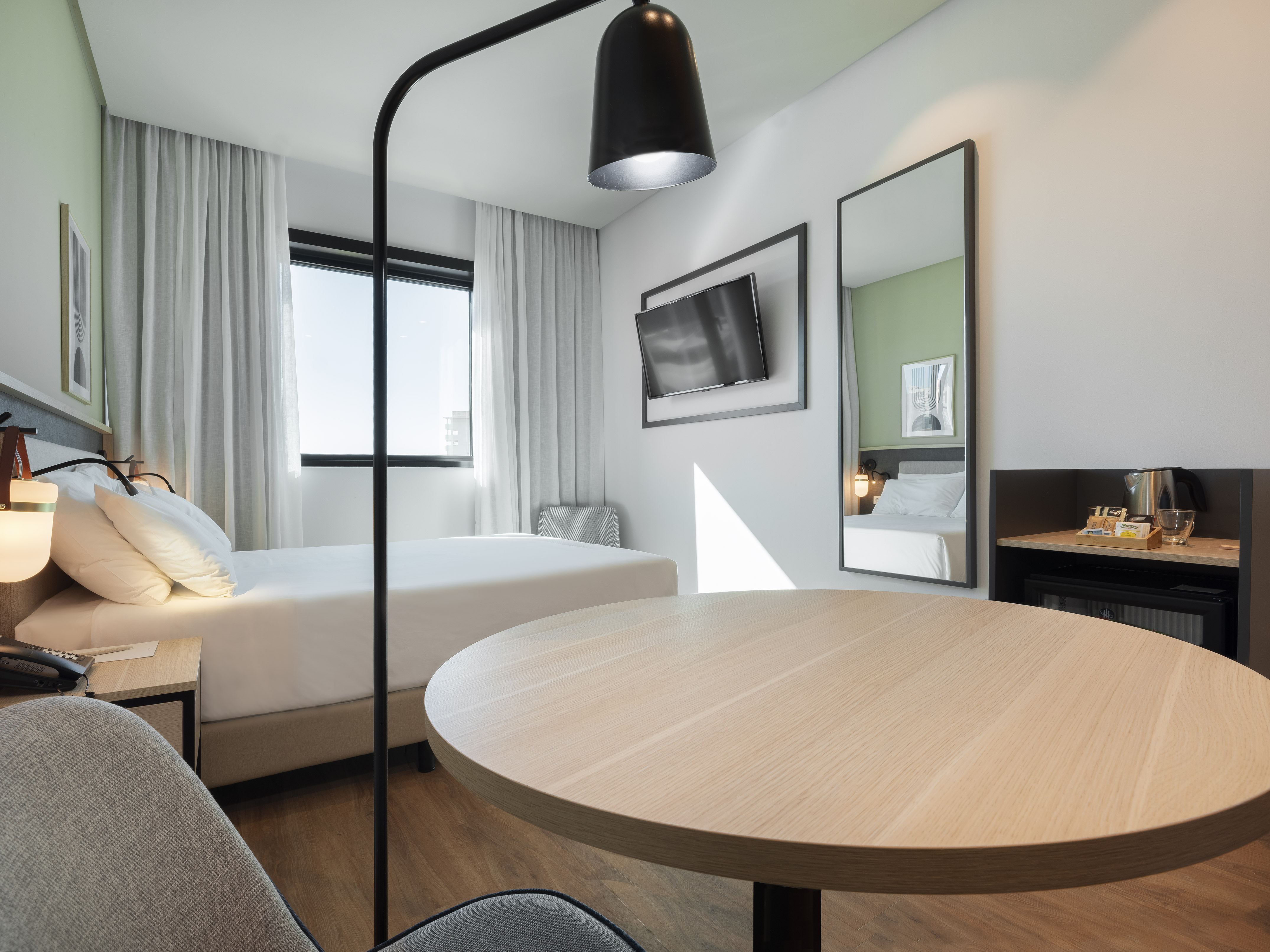 Modern hotel room with bed, round table, TV, large window, and mirror.