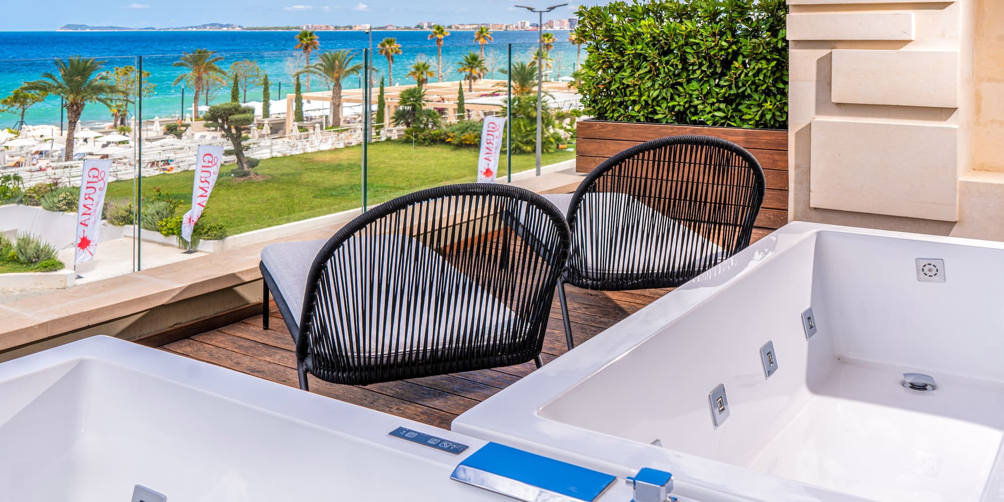 Resort balcony with two outdoor jacuzzis, chairs, beach, and blue sea view.